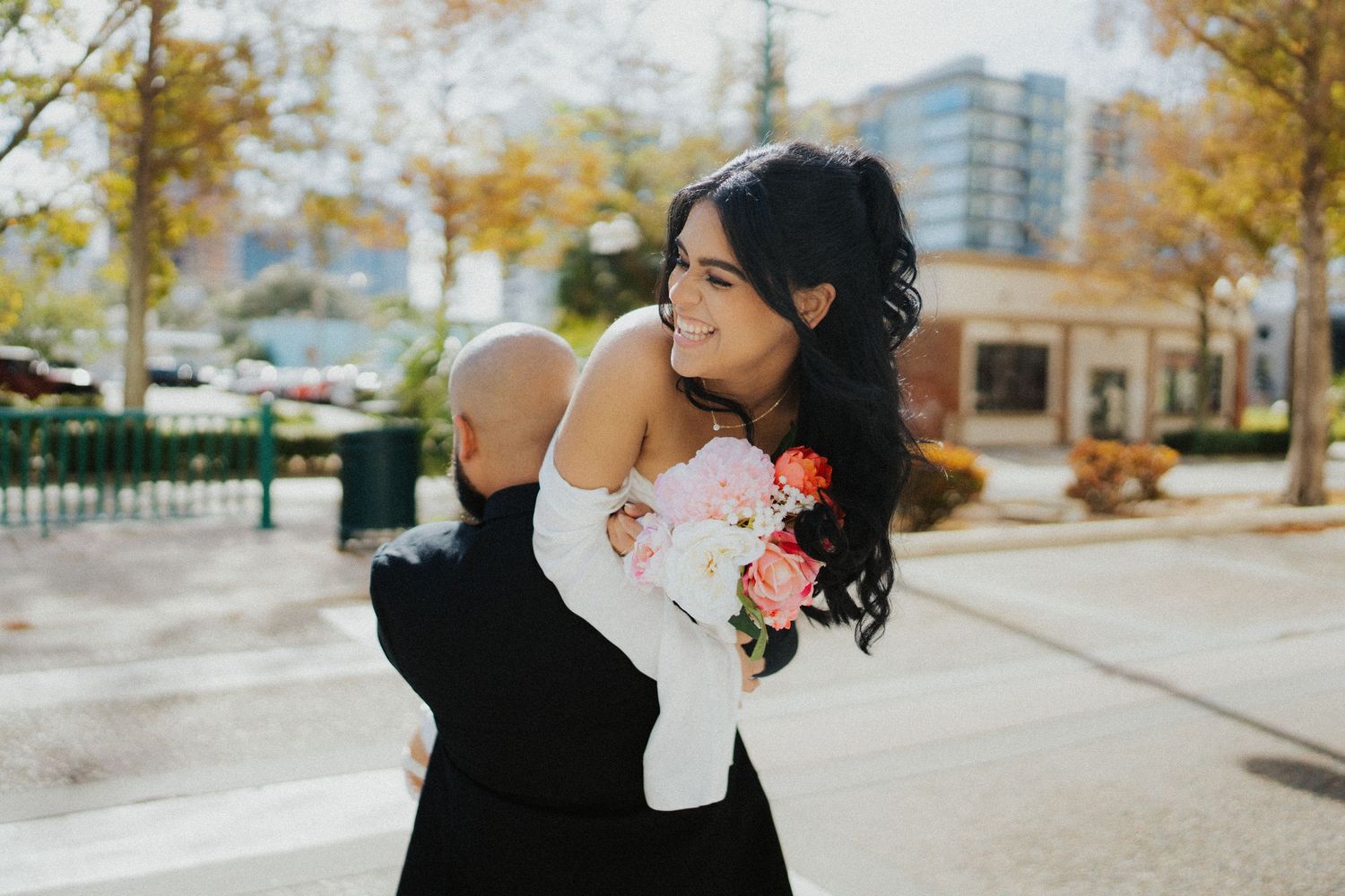 Bride in white off-shoulder gown holding pink bouquet smiles over shoulder on a city street in autumn.