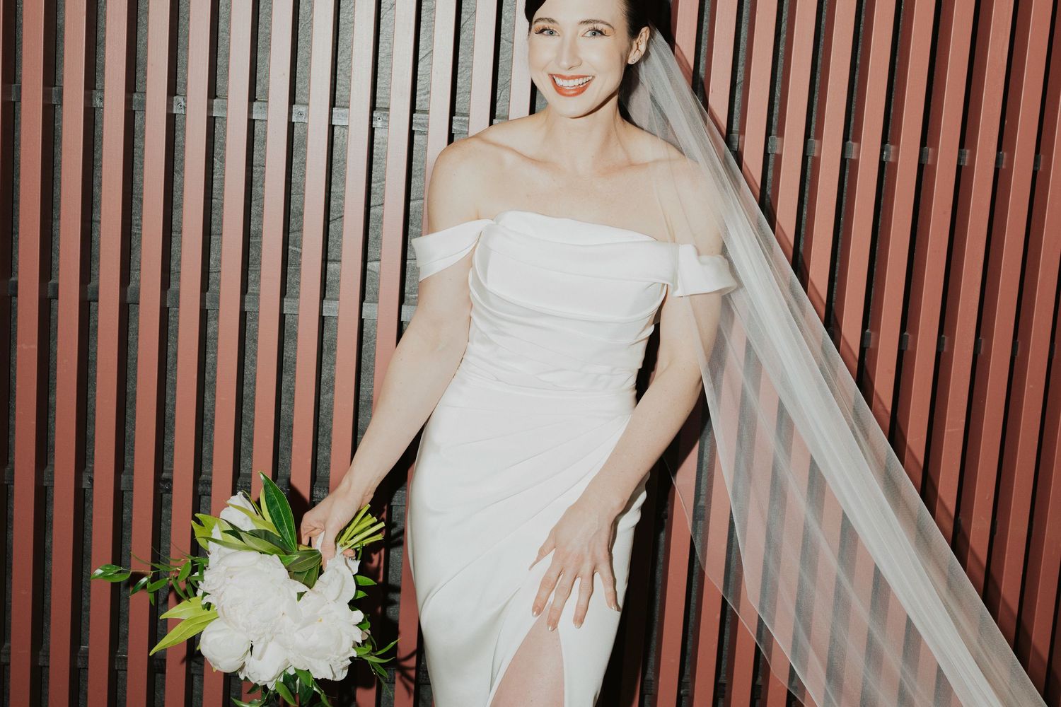 A bride in an elegant off-shoulder white wedding dress holds a white peony bouquet against a slatted red wall.