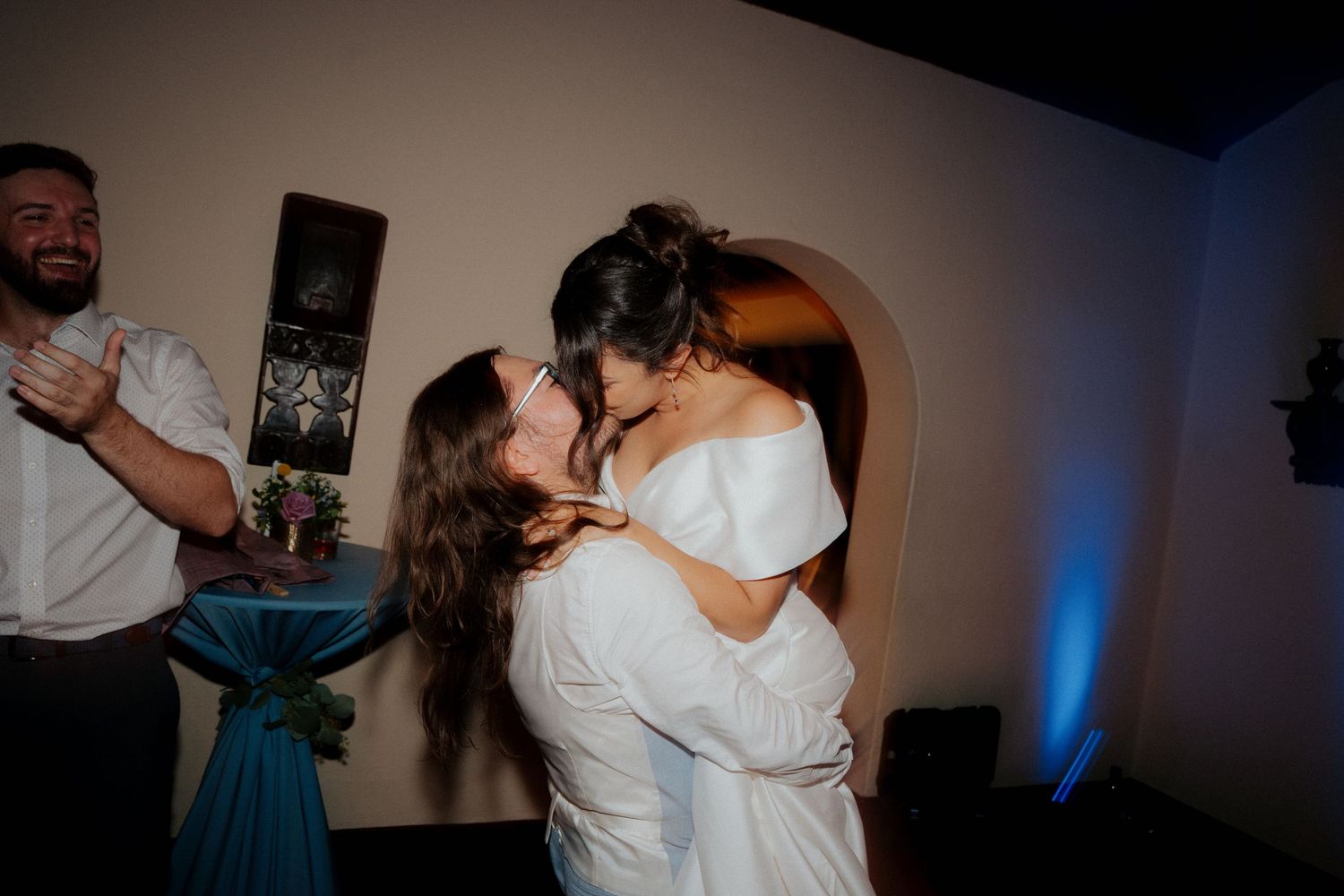 A couple in white outfits share a romantic dance moment at an indoor reception with blue accent lighting.