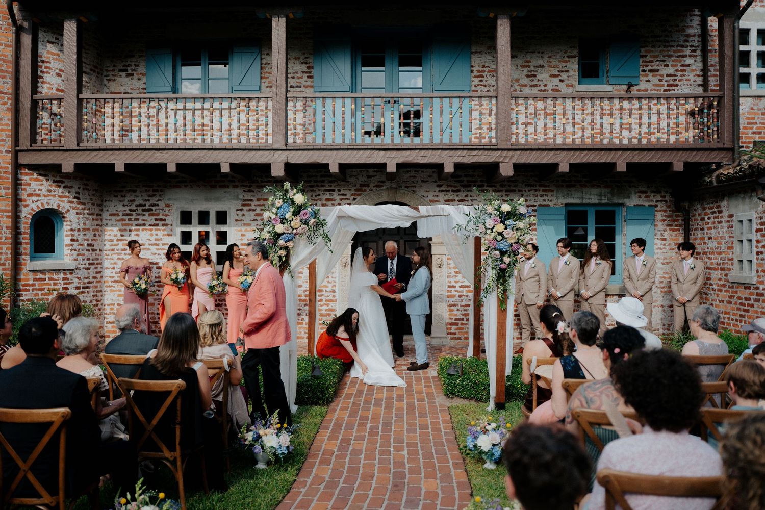 Panoramic view of a brick building exterior with guests gathered for an outdoor wedding ceremony.