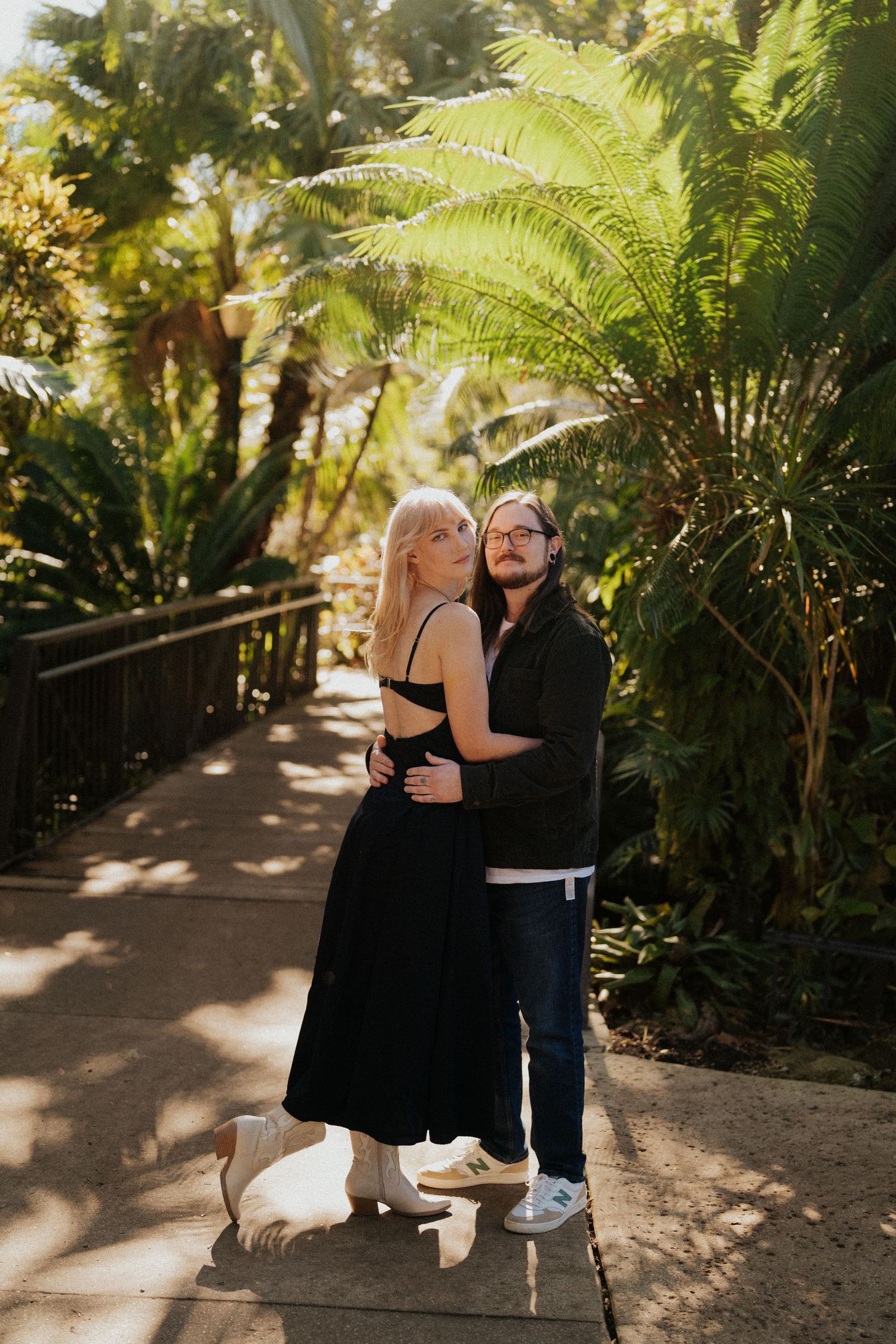 A couple embraces amid lush ferns and tropical plants on a wooden walkway.