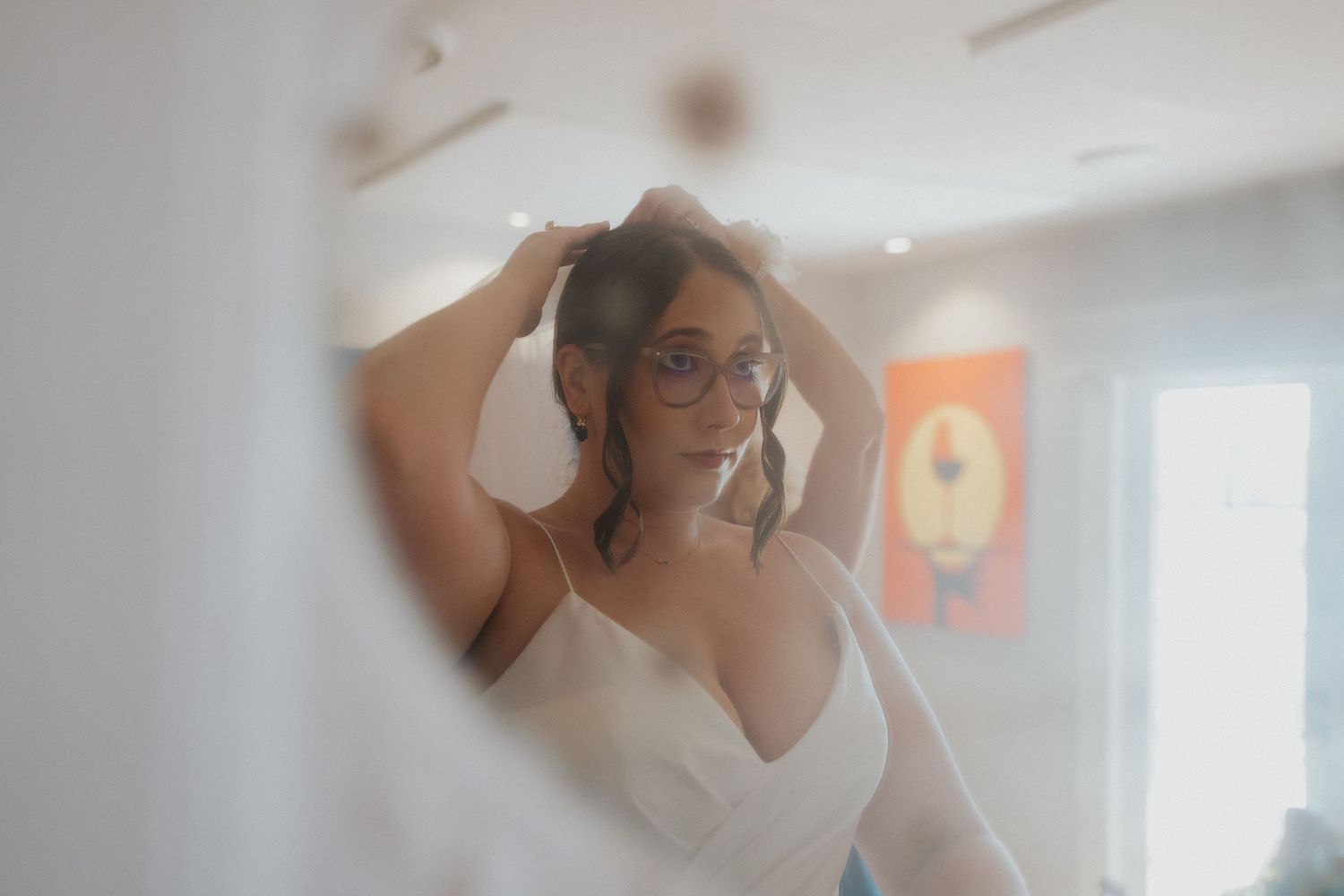 A person adjusts their hair while wearing a white dress and glasses in a softly lit indoor setting.