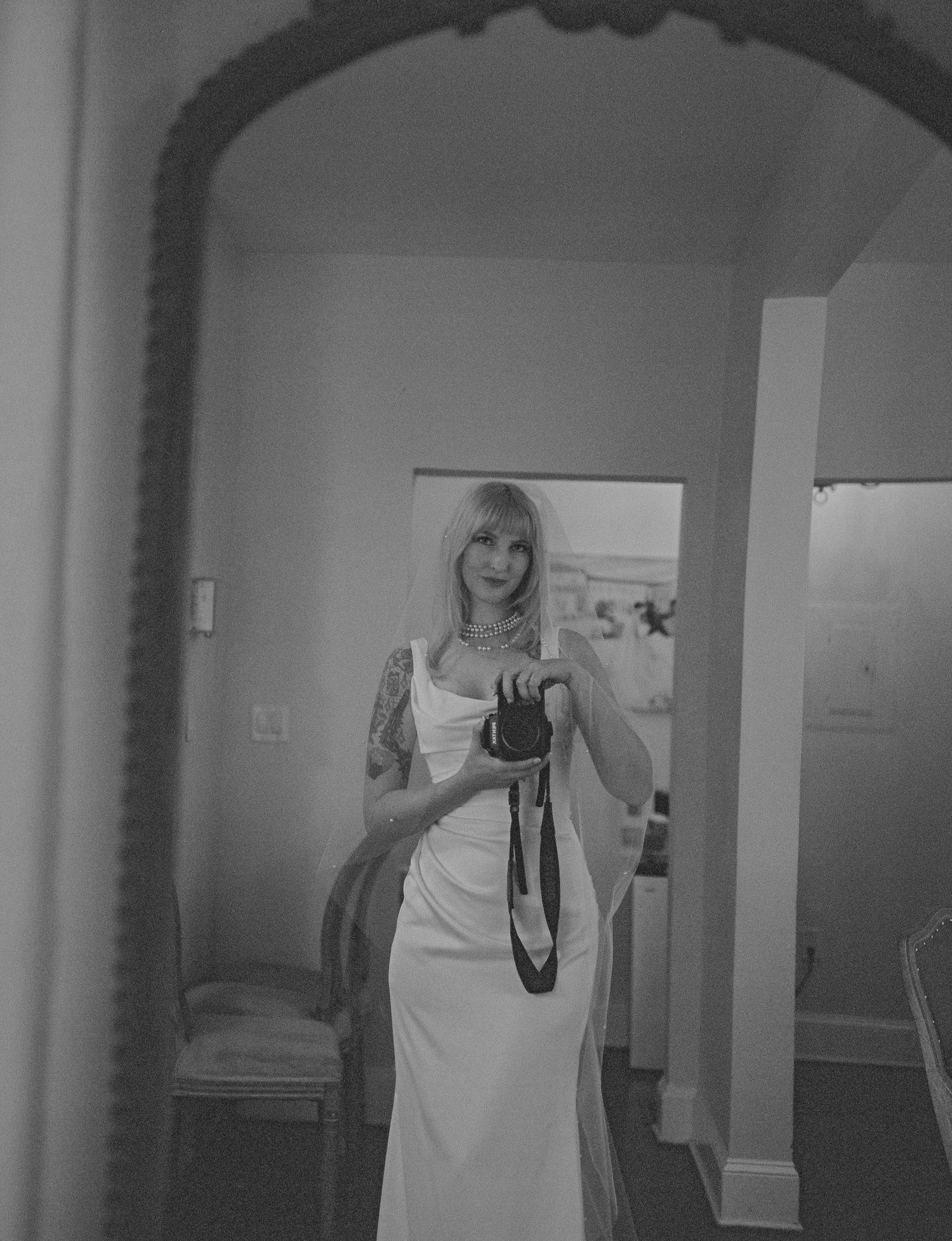Black and white mirror selfie of blonde woman in white slip dress holding a camera with tattoos.