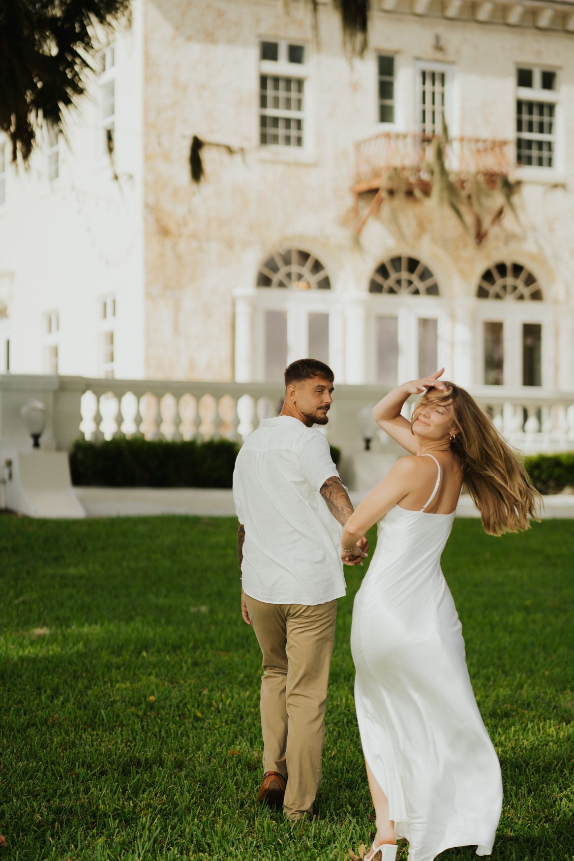 A couple in white and khaki clothing dance joyfully on a lawn in front of an elegant white mansion with arched windows.