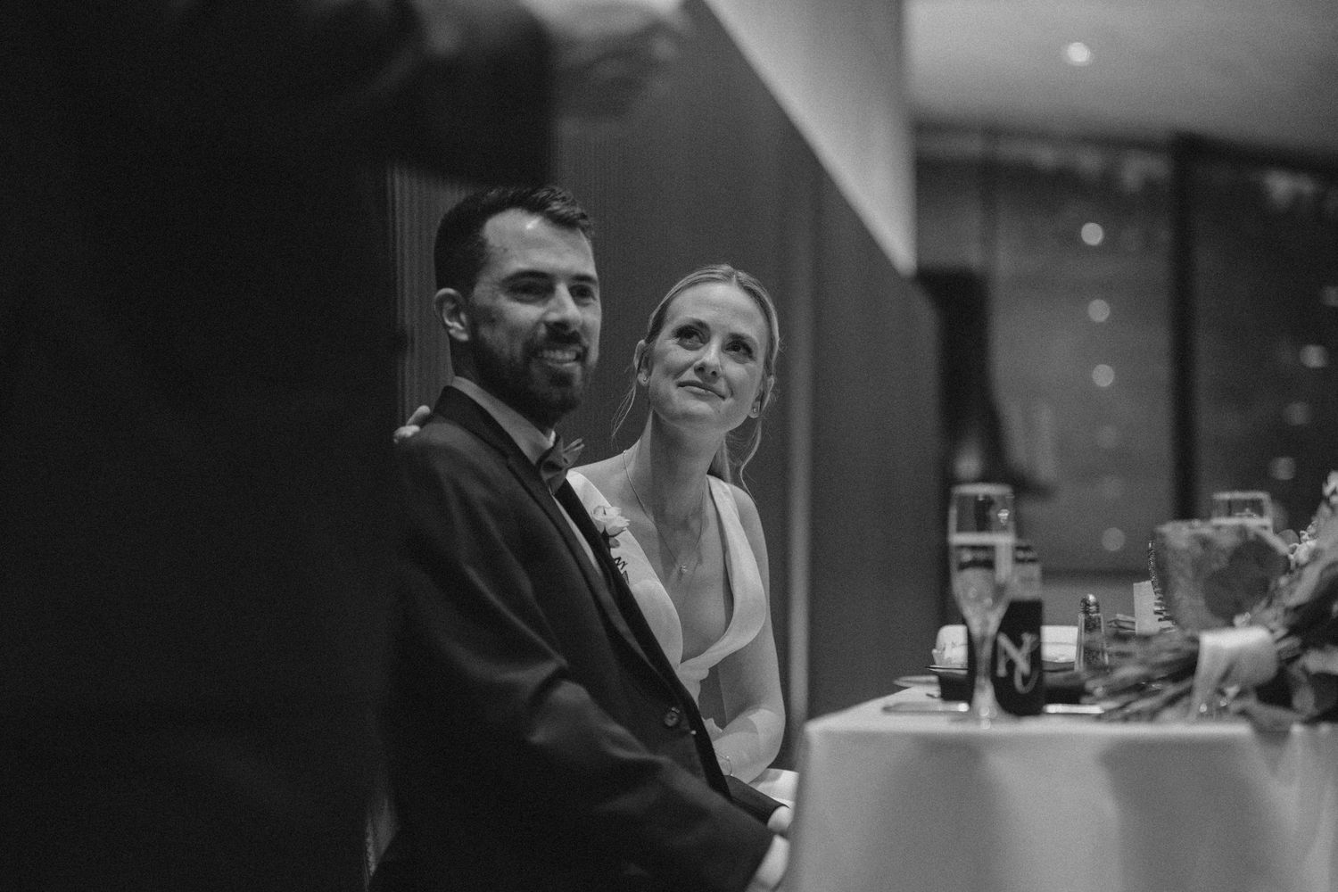 A black and white candid photo of a couple smiling together at a social gathering or event.