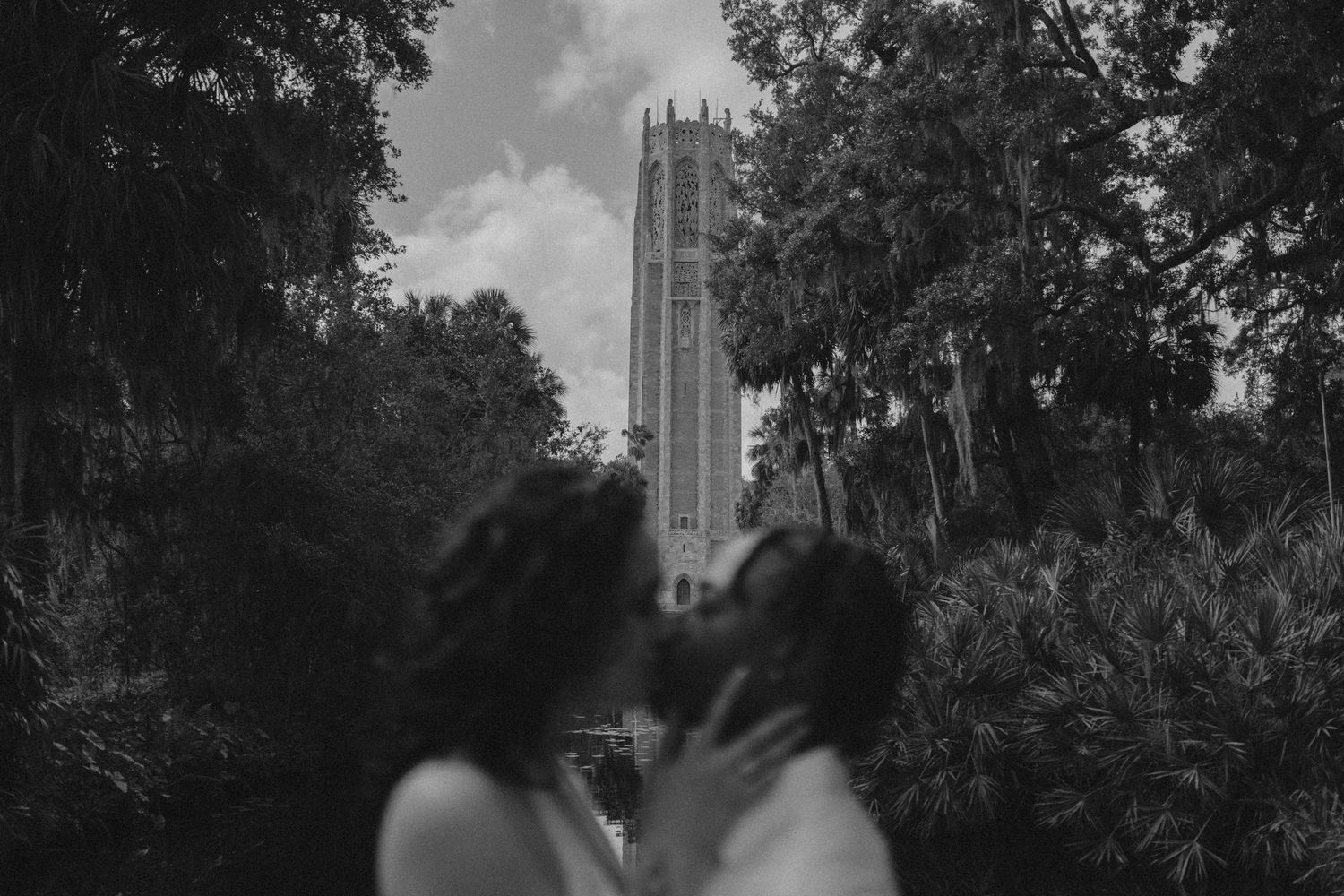 Black and white photo of couple kissing with Bok Tower rising above the trees in background.