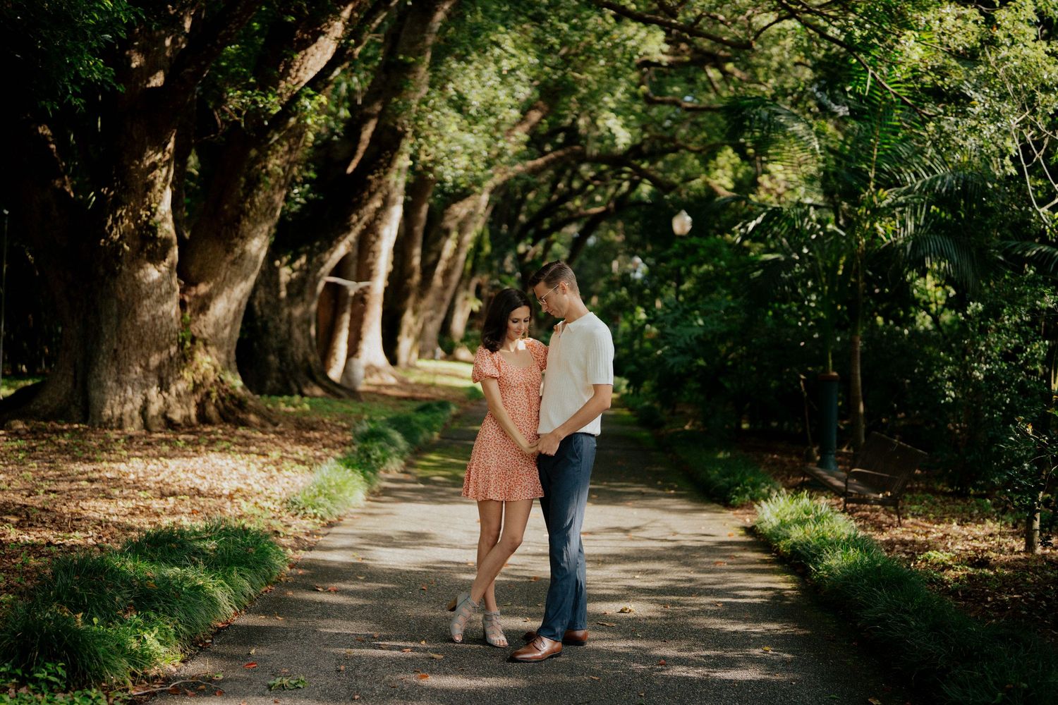 Couple embraces on a tree-lined path dappled with sunlight, woman wearing a pink floral dress.