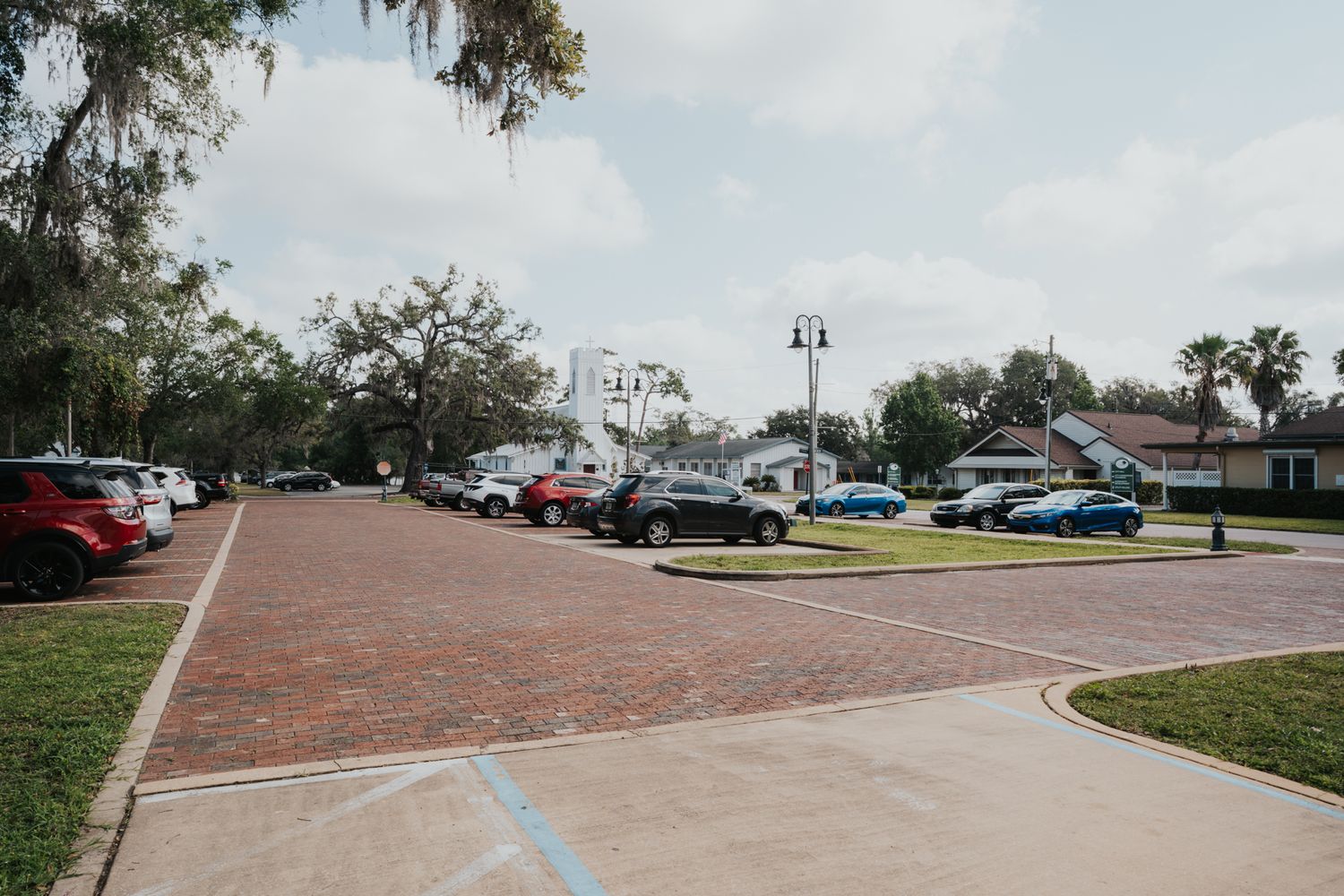 Brick paved parking lot with cars and lamp posts surrounded by trees and greenery.