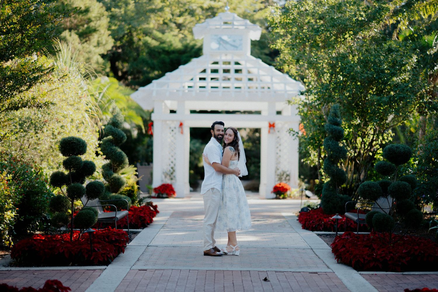 White gazebo surrounded by landscaped gardens and red flowers creates an elegant outdoor setting.