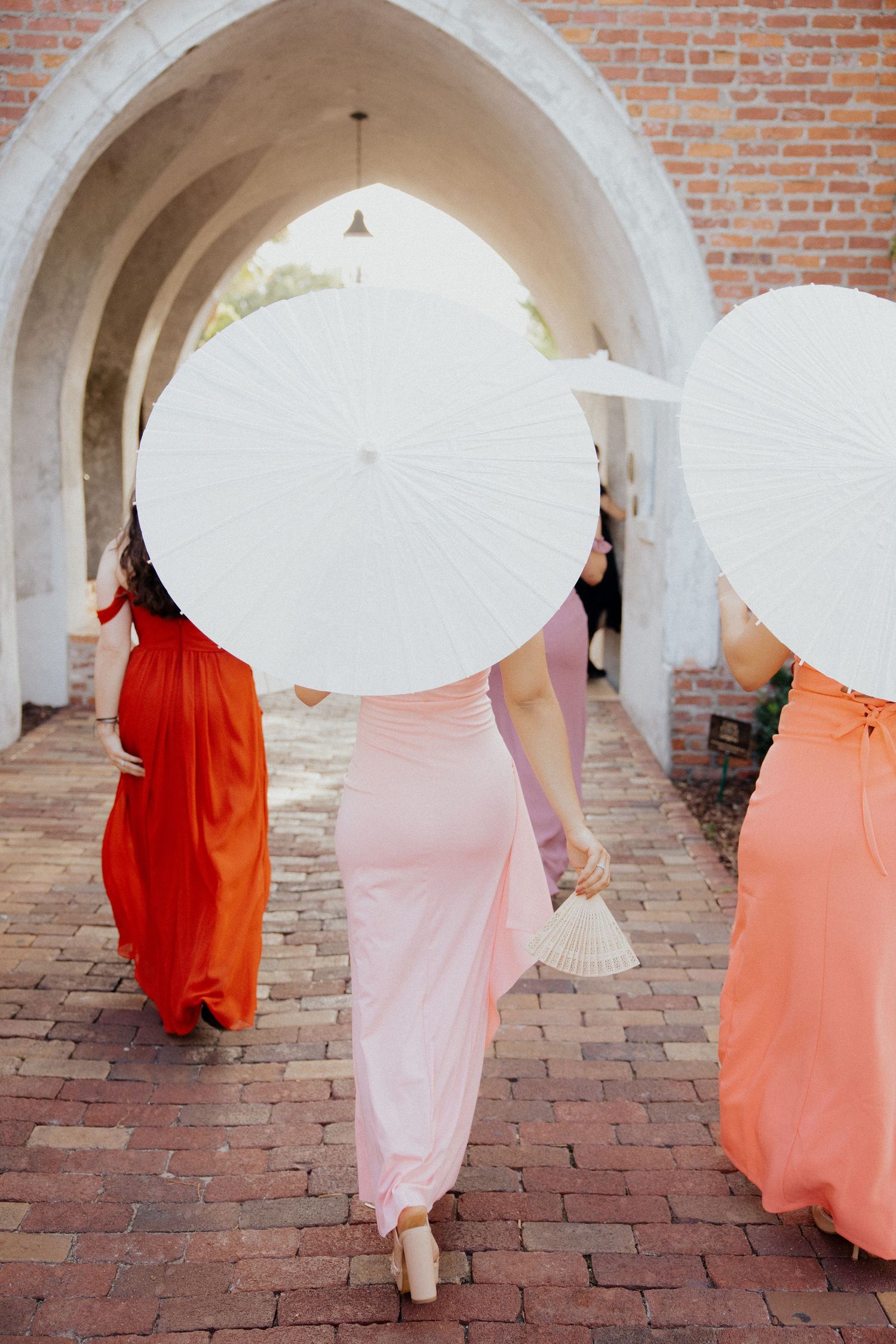 Wedding party in coral and pink dresses walks under brick archways on red cobblestone.