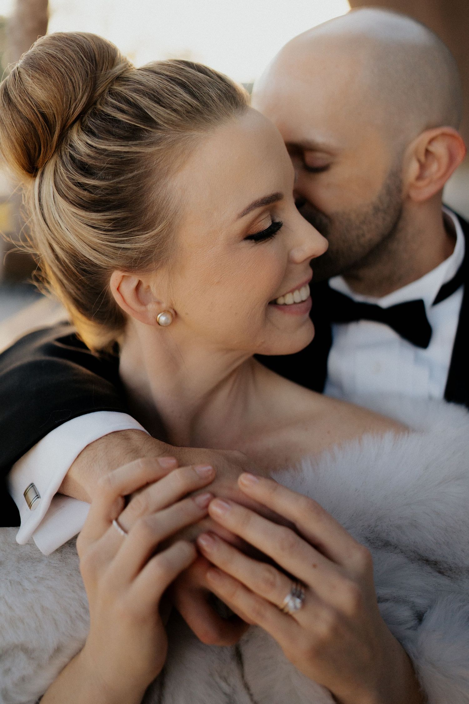 Smiling bride with updo and pearl earring held close by groom in black tuxedo.