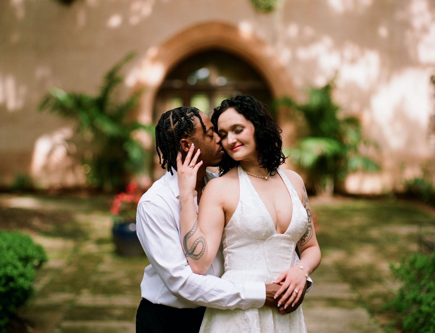 Couple sharing a tender kiss in a wedding portrait against a rustic terracotta wall with lush greenery.