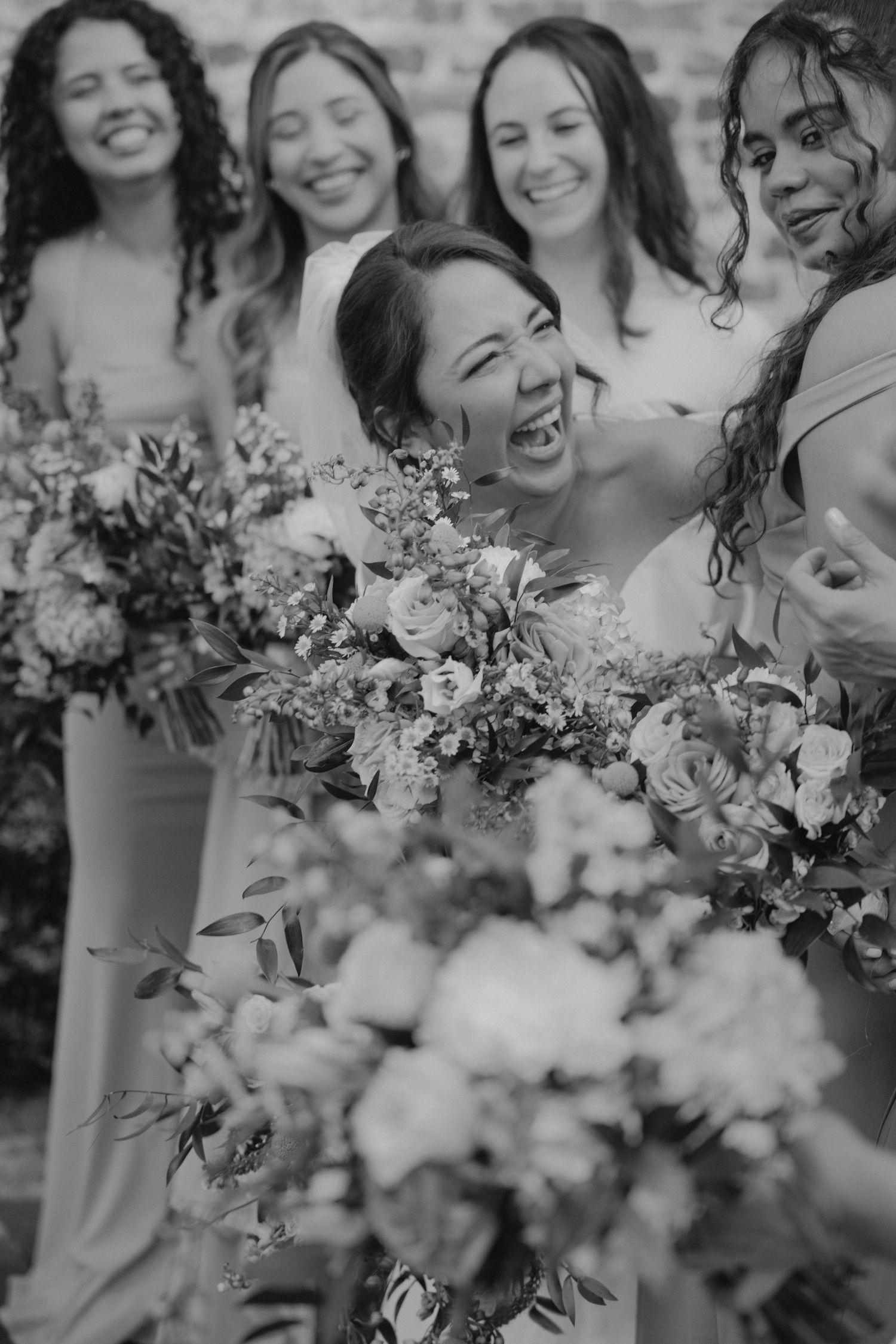 Joyful wedding party holding delicate floral bouquets against a rustic brick wall.