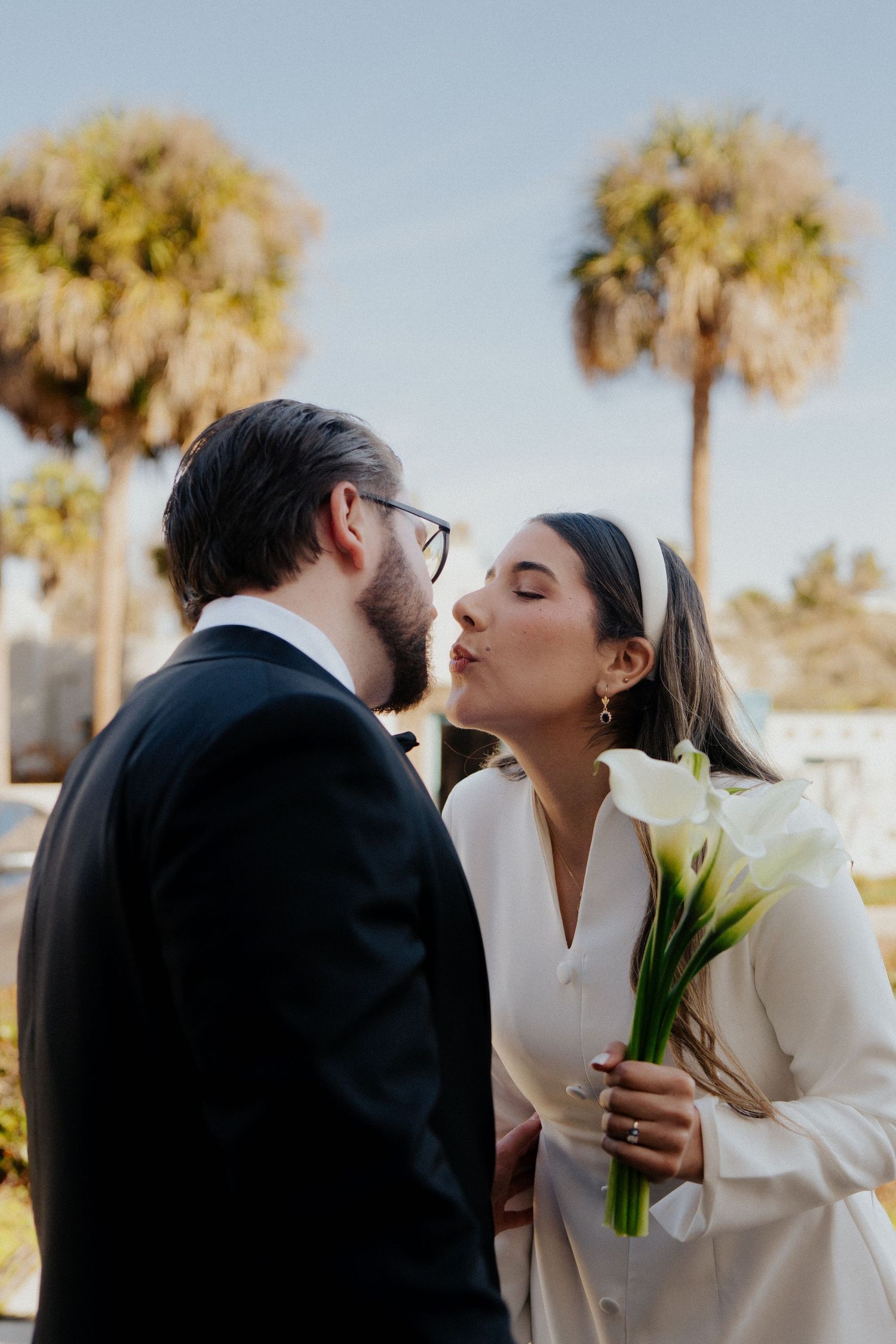 A couple shares a tender moment outdoors while the bride holds white calla lilies against a palm tree backdrop.