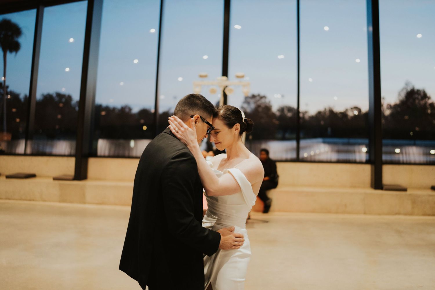 A couple shares a tender moment on a dance floor with large windows and natural light streaming in.