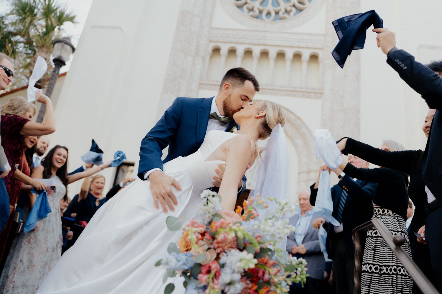 A joyful wedding celebration outside a church with guests throwing graduation caps as the couple shares a romantic moment.