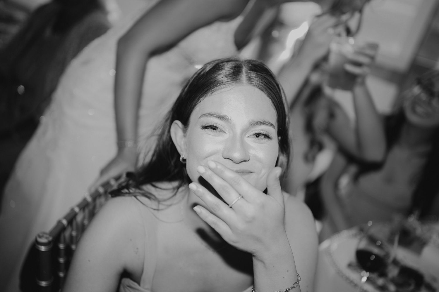 Black and white candid photo of a smiling young woman covering her mouth at a wedding reception.