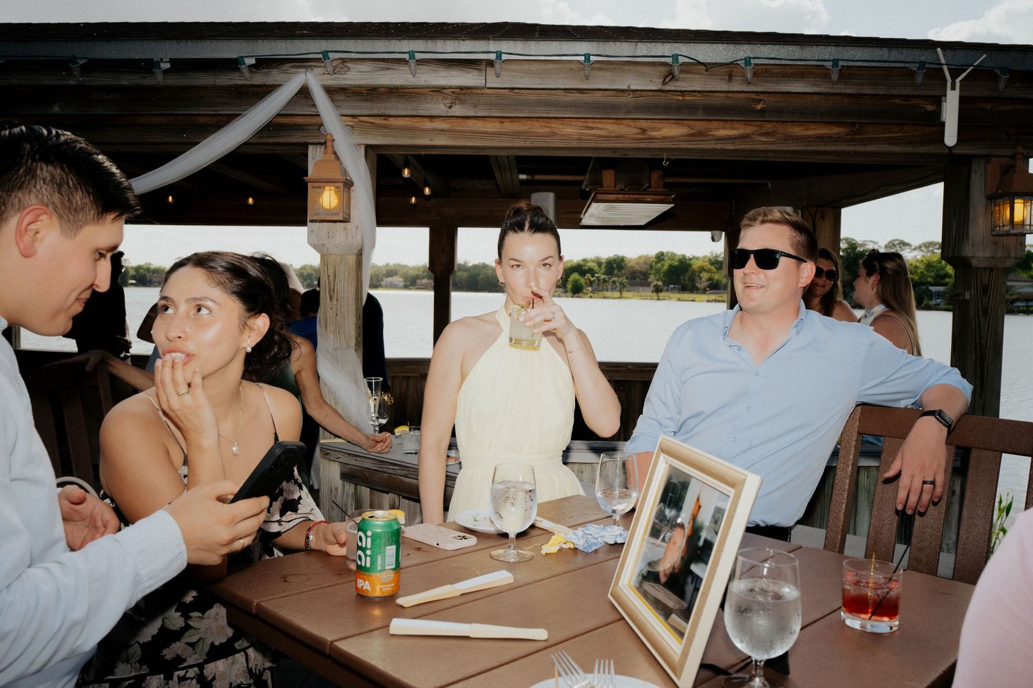 Wedding reception guests seated at table with framed photo centerpiece in open-air venue.