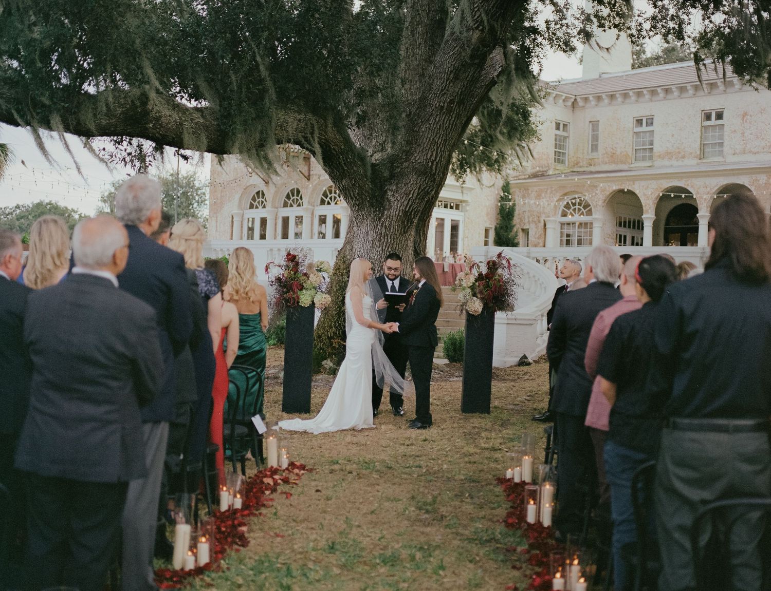 An outdoor wedding ceremony under a large tree with guests gathered around candle-lined aisle at a historic venue.