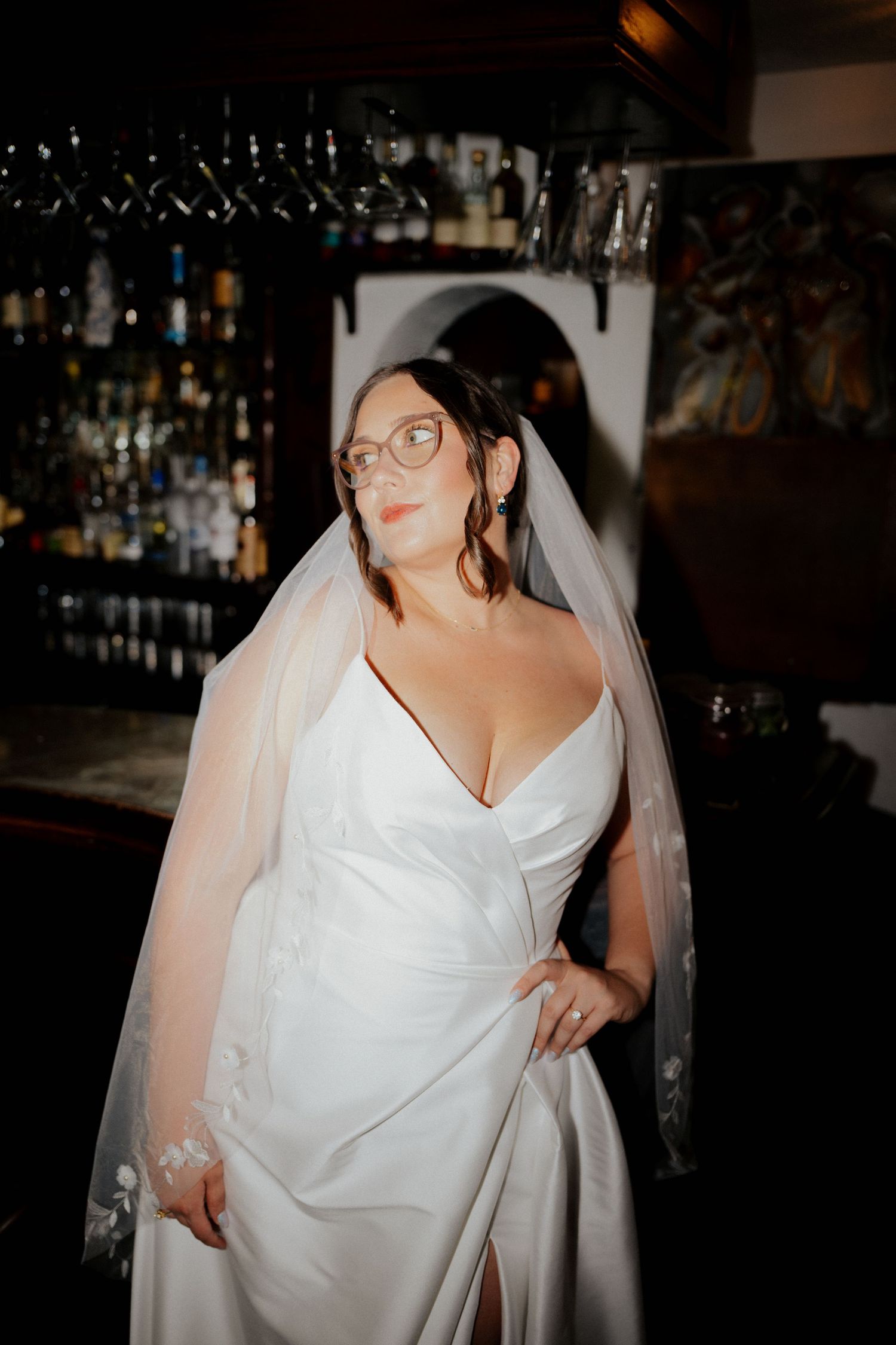 A bride in a classic white wedding gown and veil poses at a enzos on the lake bar with glasses and bottles in the background.