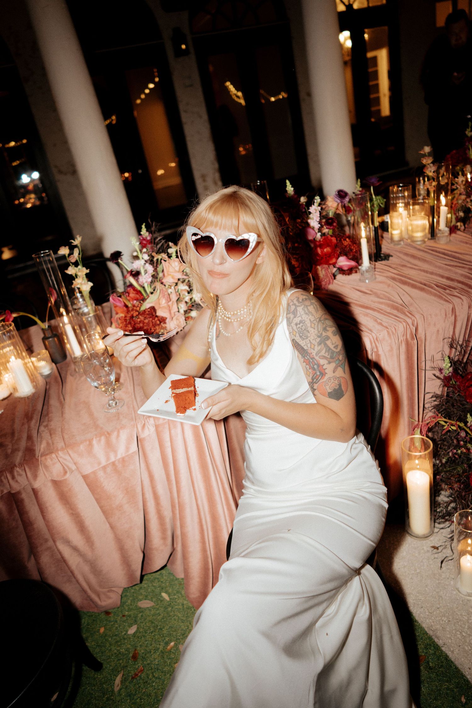 Bride in white gown and heart-shaped sunglasses sits at candlelit reception table with pink floral arrangements.