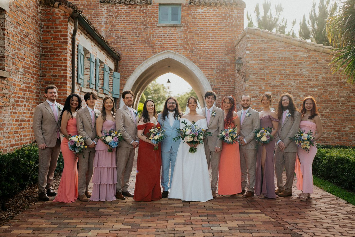 Wedding party members in coordinating pink and mauve dresses pose together against rustic brick walls at an elegant venue.