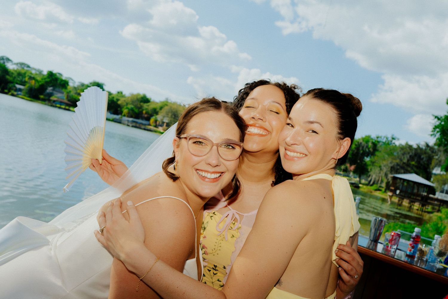 Three women embracing and smiling together on a boat with a scenic lake backdrop.