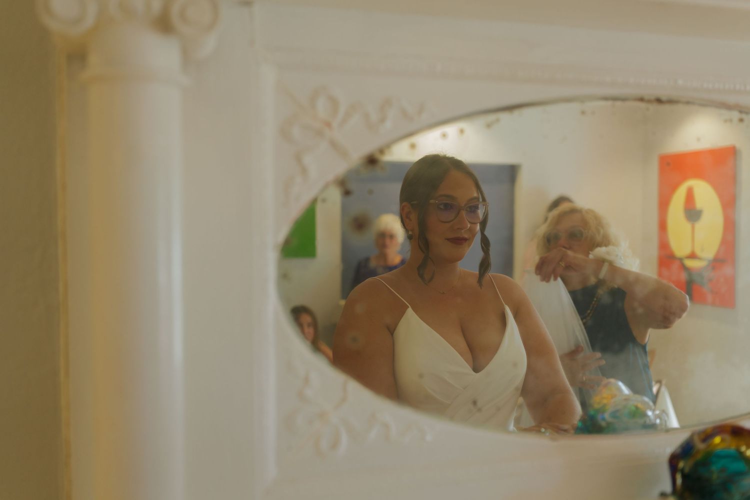 Woman in white dress admiring her reflection in an ornate oval mirror, with colorful artwork visible in the background.