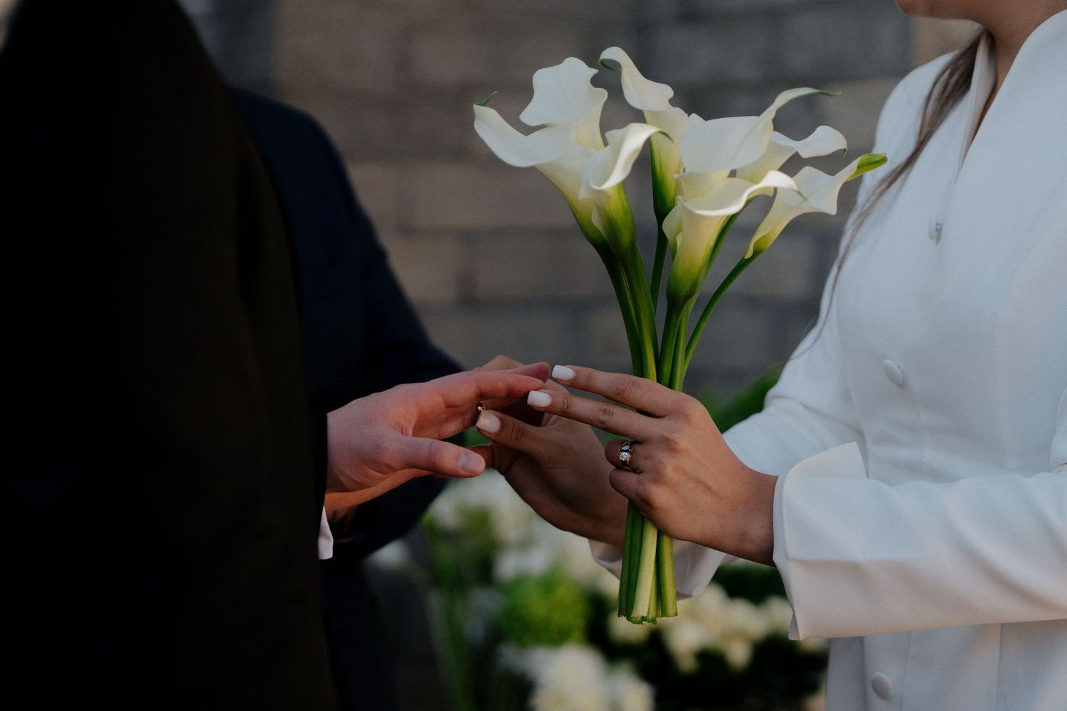 A close-up shot of hands exchanging rings during a wedding ceremony with white calla lilies in the background.