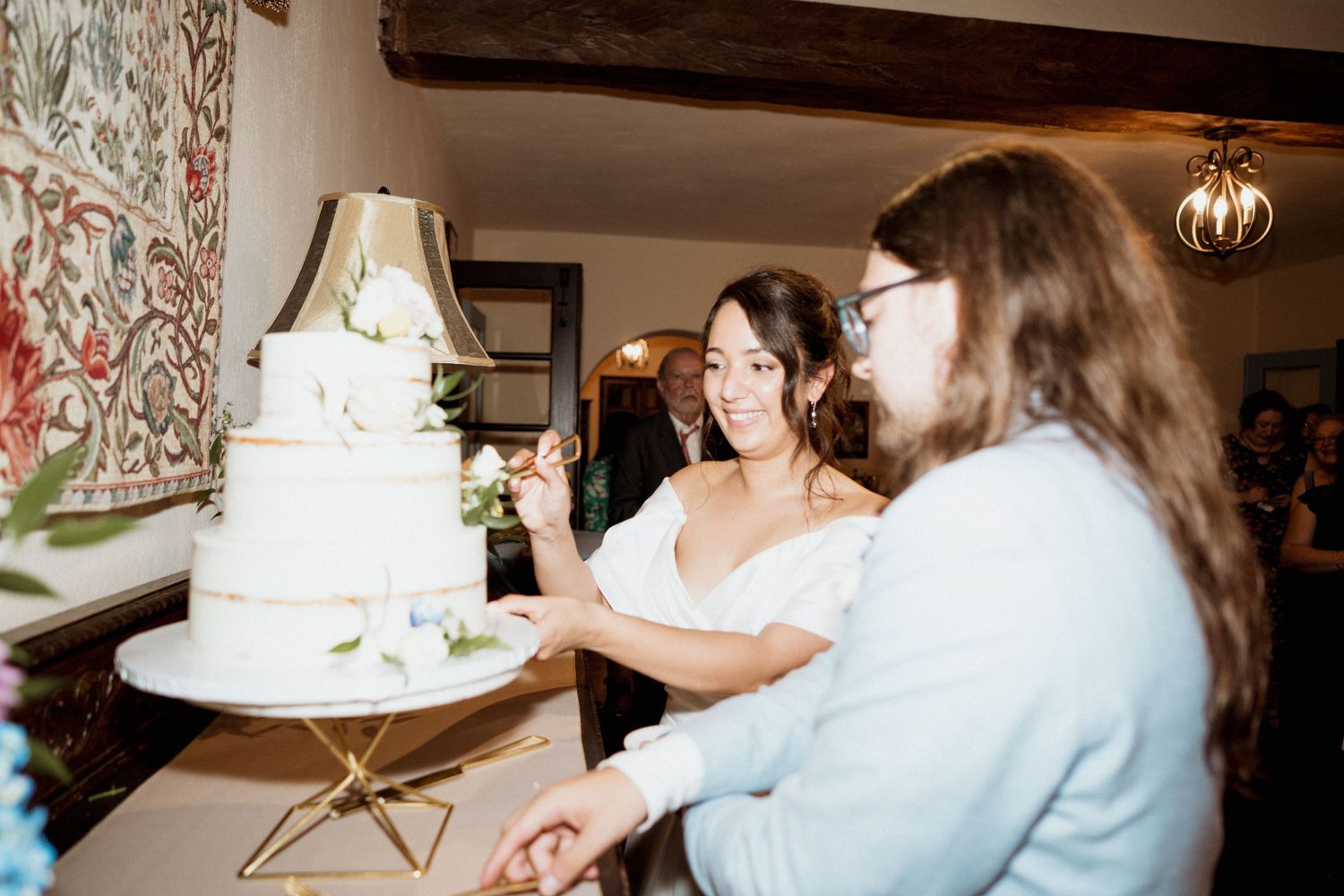 A couple cuts into their elegant white tiered wedding cake decorated with delicate floral accents at their reception.
