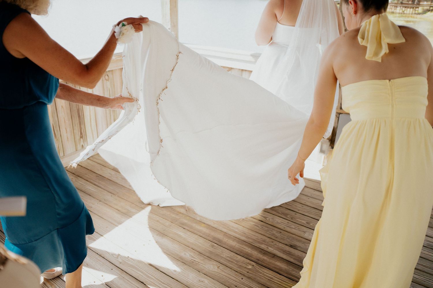 Bridesmaids helping bride with white wedding dress train on wooden deck outdoor venue.