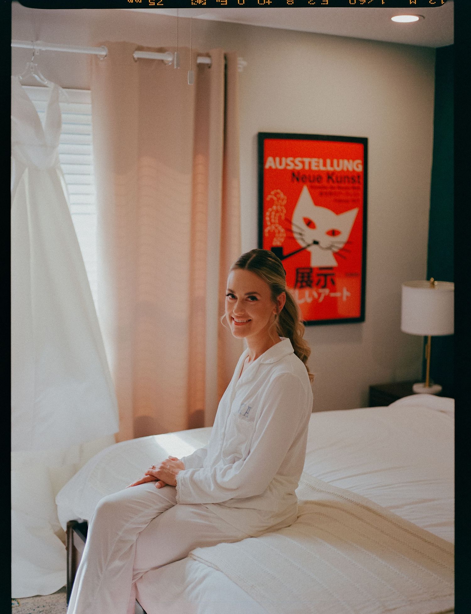 Smiling bride in white robe sits on bed near hanging wedding dress in hotel room.