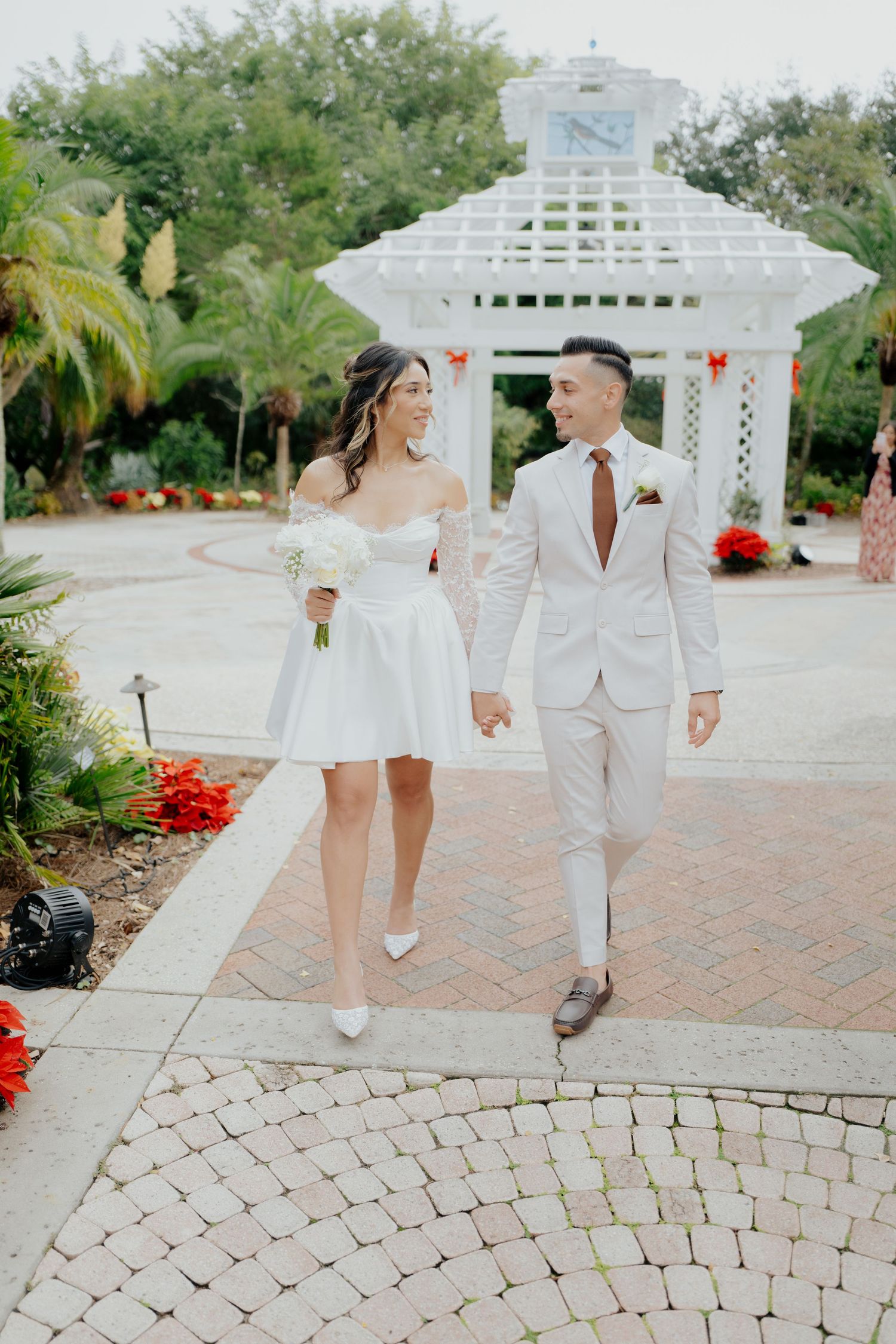 A couple in matching white outfits walks hand in hand along a curved brick pathway by a garden gazebo.