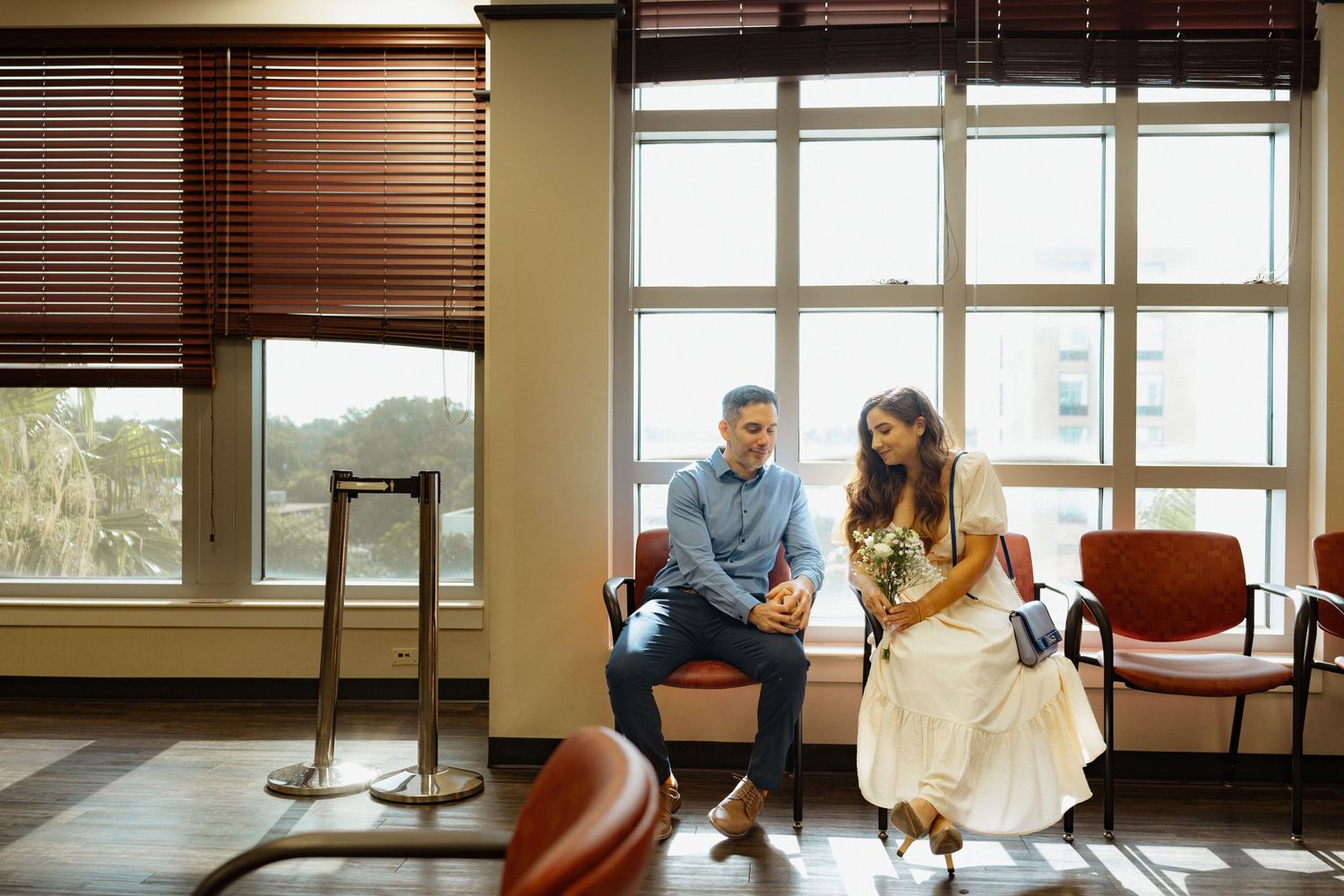 A couple sits together on chairs by a window with wooden blinds in an indoor setting.