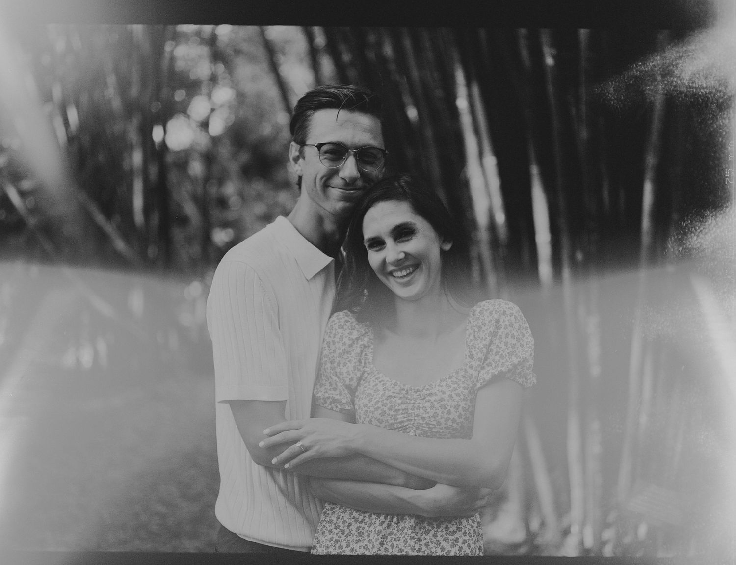 A black and white portrait of a couple embracing outdoors with trees in the background.