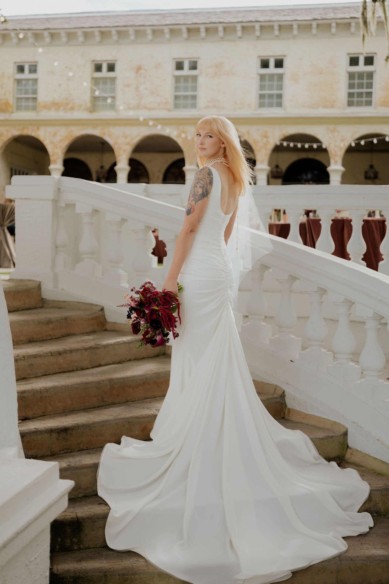 Bride in elegant white gown with train poses on stone staircase holding deep red bouquet at Bella Cosa in Lake Wales