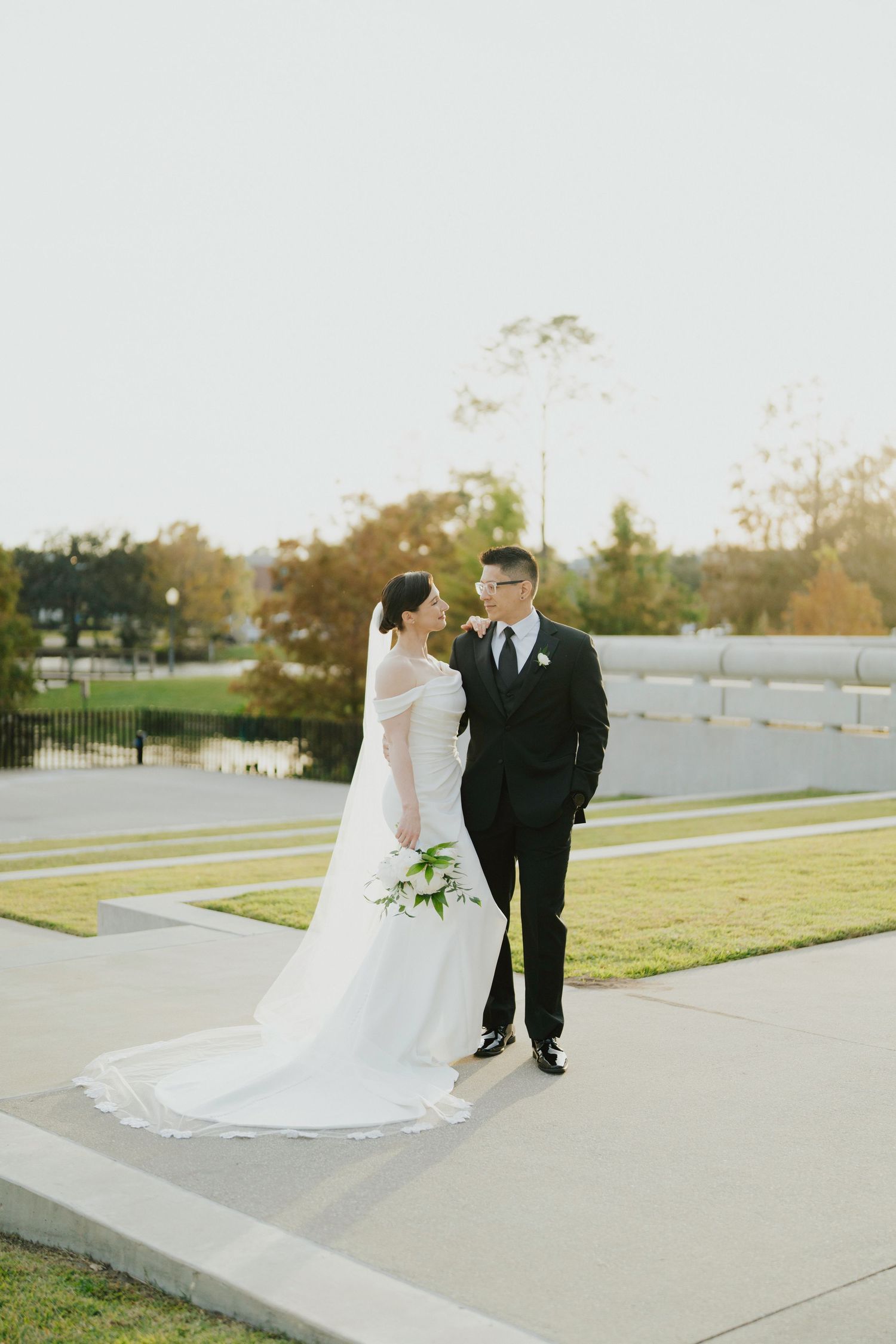Bride and groom in classic black tuxedo walking on pathway with lush park backdrop.