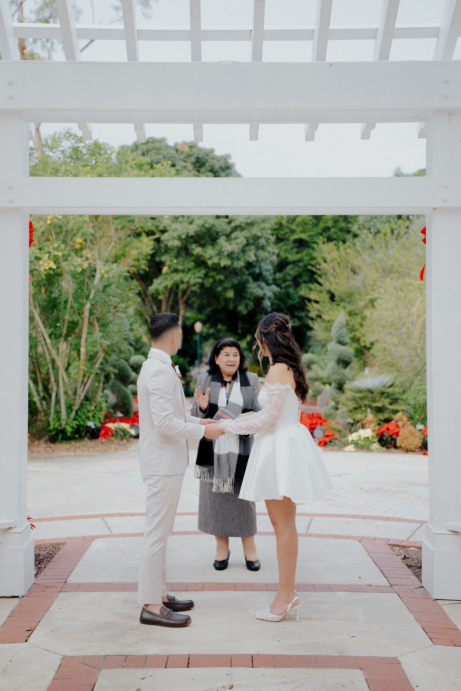 A couple shares an intimate moment on a white gazebo deck surrounded by lush greenery and red flowers.