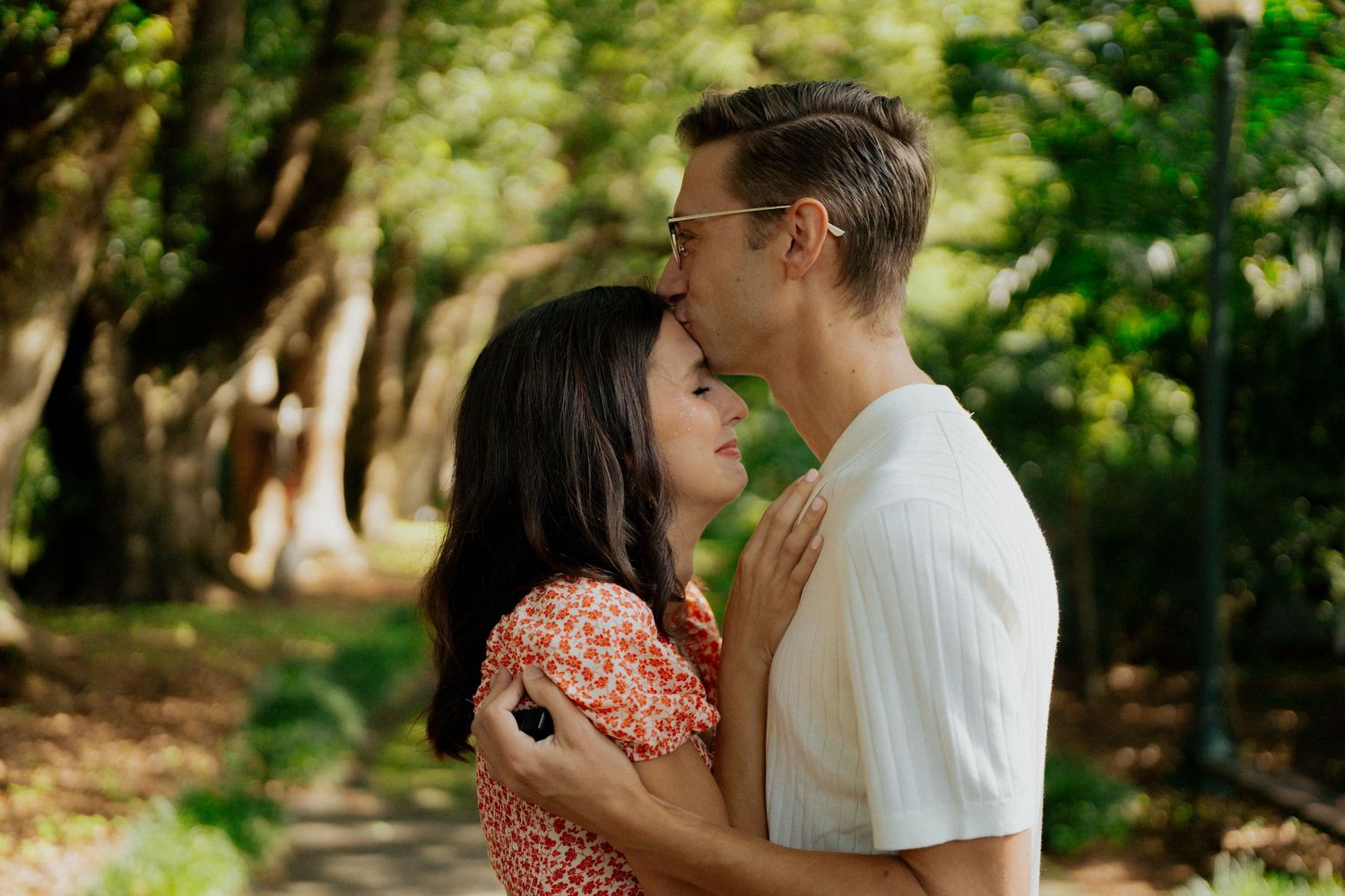 A couple embraces and laughs together while walking along a sunlit wooded path.