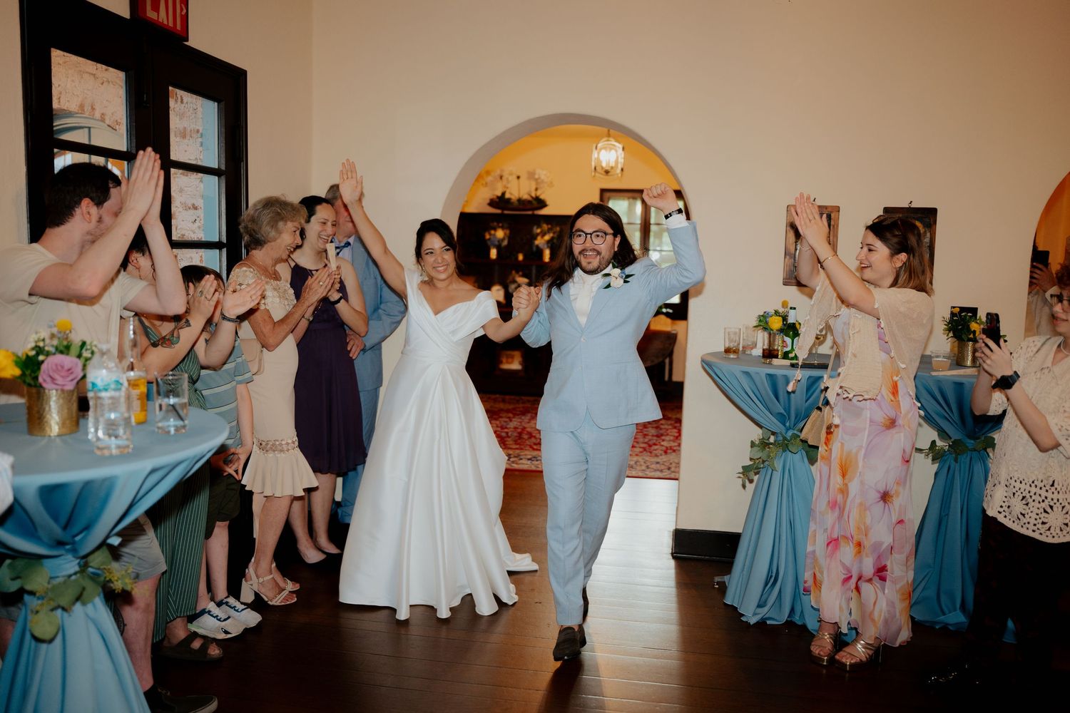 Wedding party members in blue and floral dresses dance together in celebration at an indoor reception.