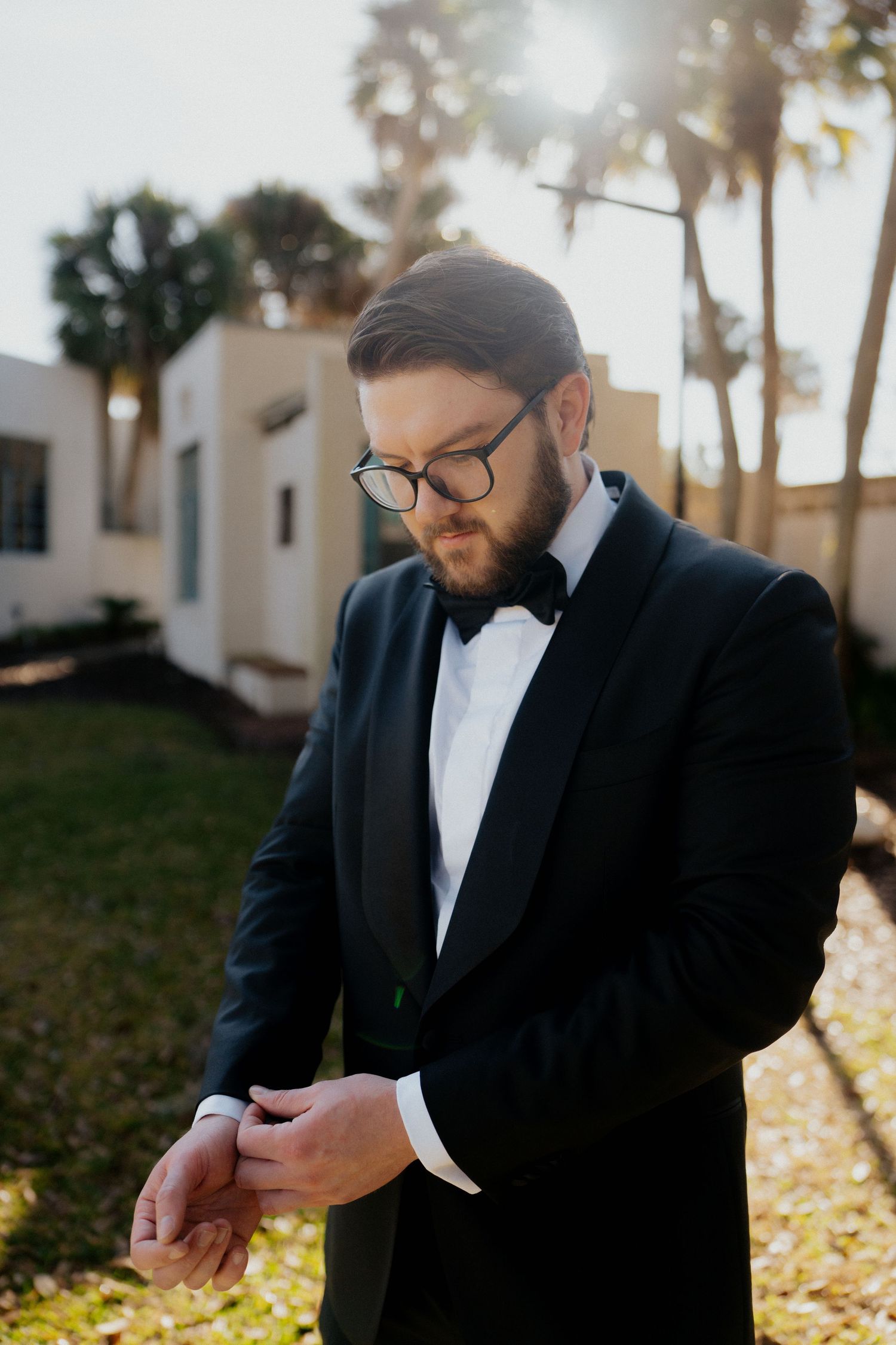 A groom in a black tuxedo and bow tie checks his watch in warm sunlight with palm trees in the background.