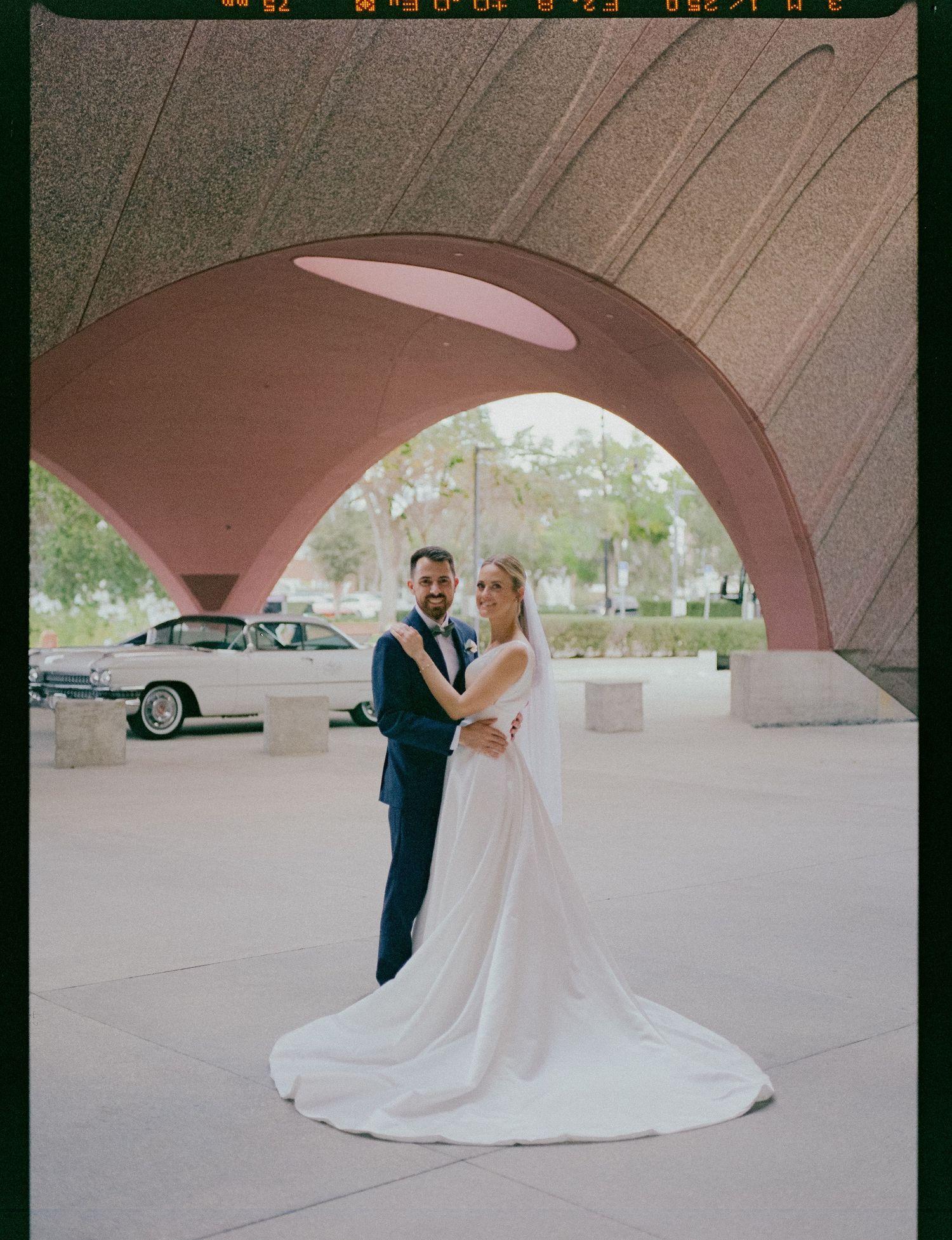 A couple poses under a curved pink archway on their wedding day, with a classic car visible in the background.