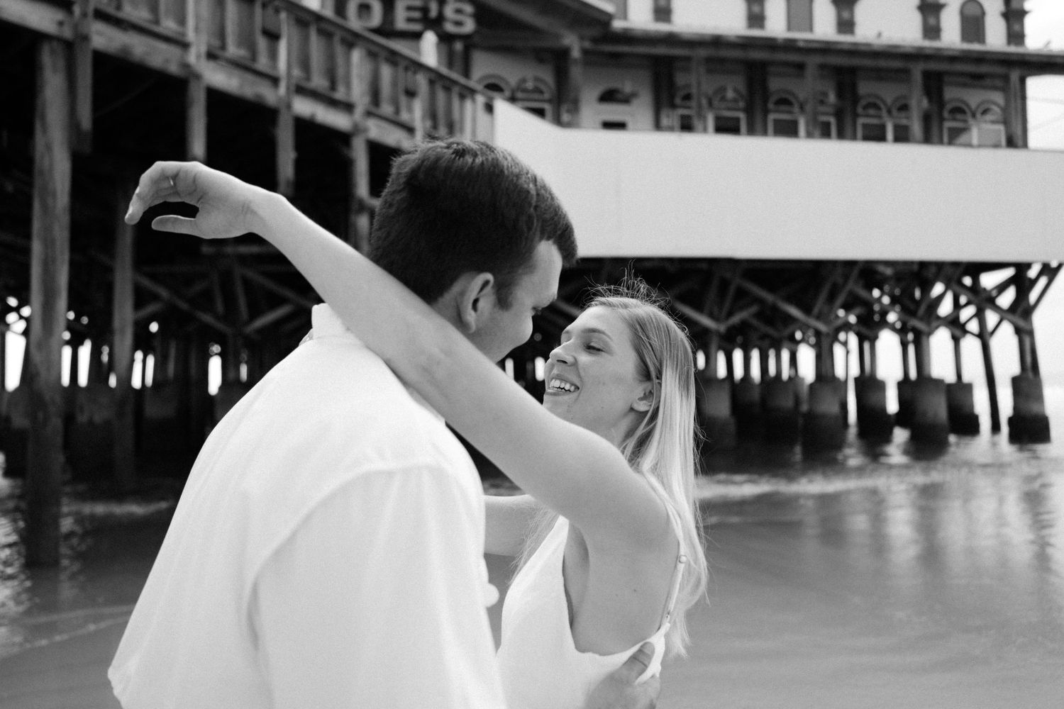 A tender embrace captured in black and white beneath a classic beachside pier.