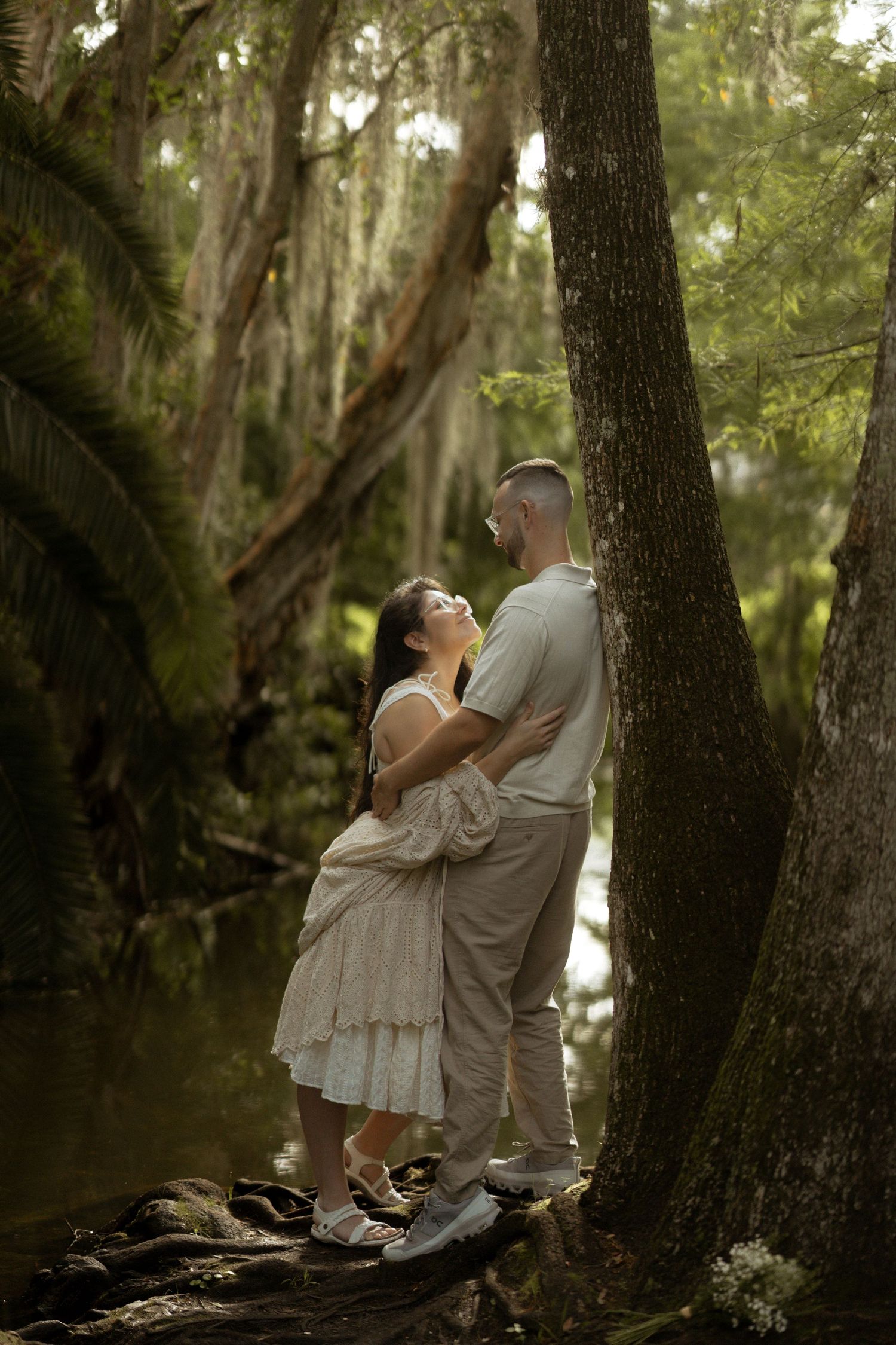 Couple embracing by mossy tree trunk near reflective water in lush green forest setting.