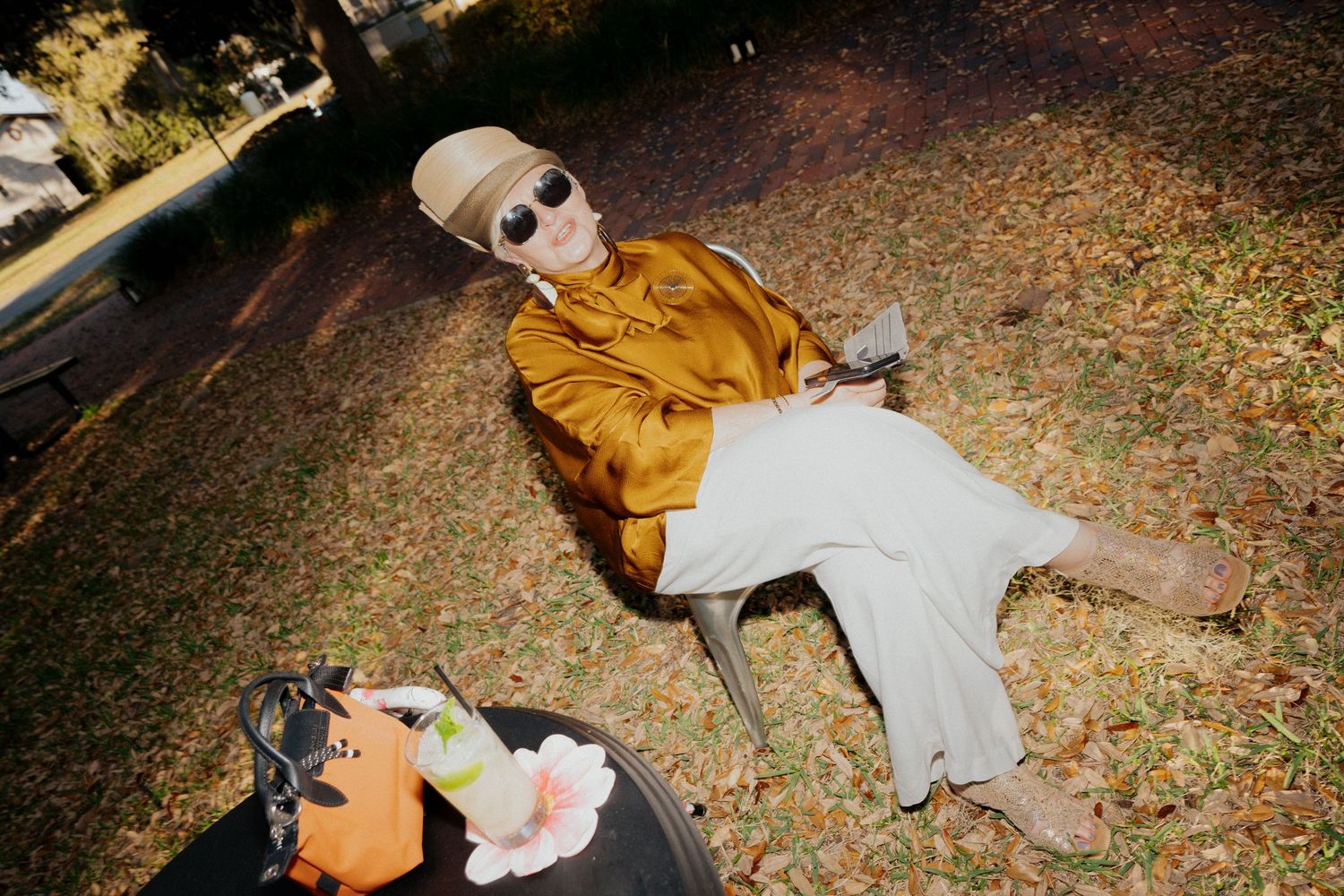 Person in golden jacket and white pants relaxing on grass with sunglasses and hat, autumn leaves scattered around.