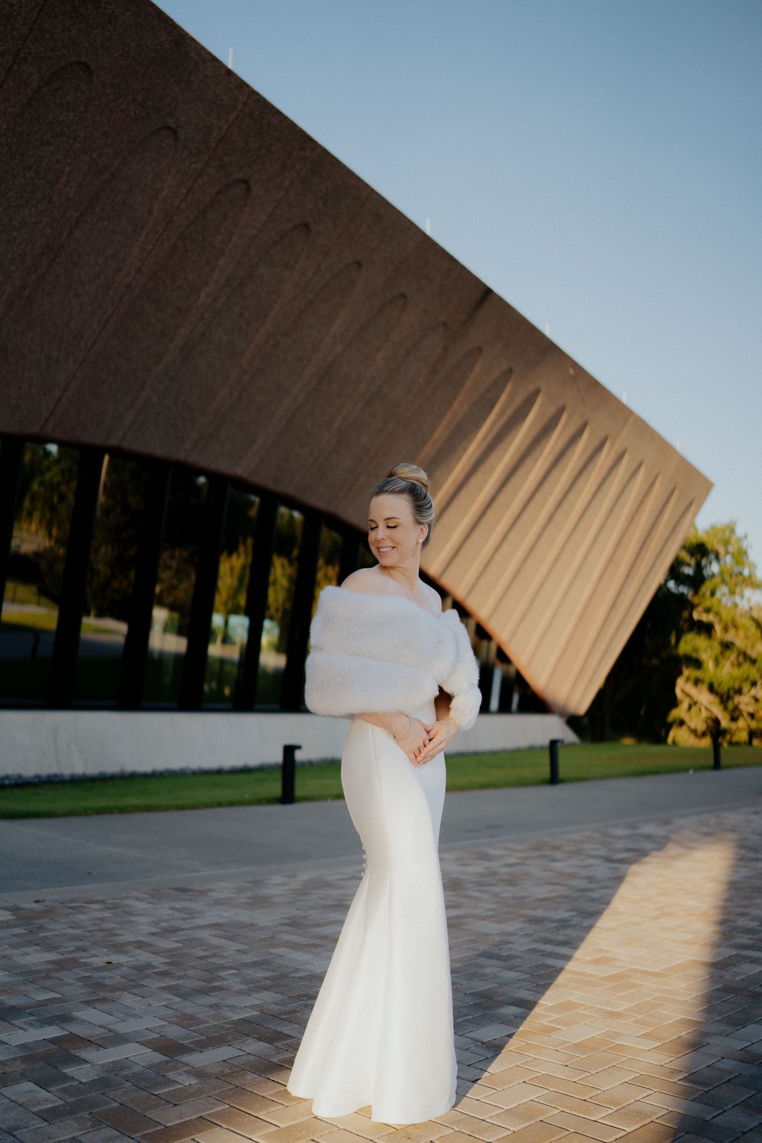 Elegant bride in white mermaid gown and fur stole poses before modern architectural building.