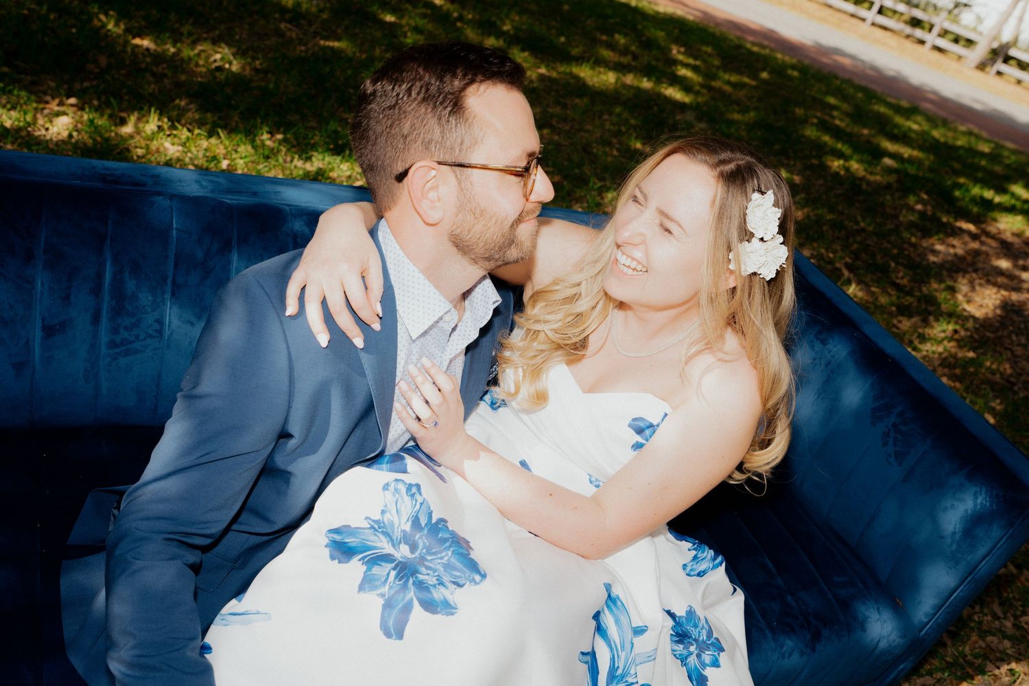 Smiling bride and groom sitting together on blue velvet outdoor sofa at wedding.