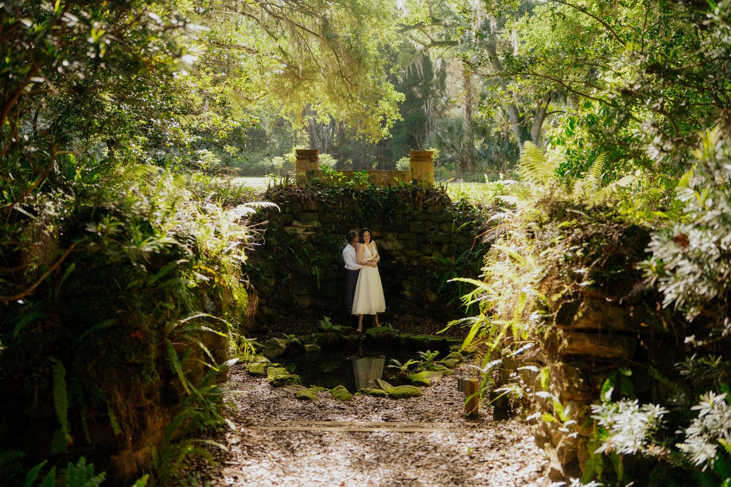 Couple embracing in a romantic lush green garden pathway framed by tropical foliage and stone walls.