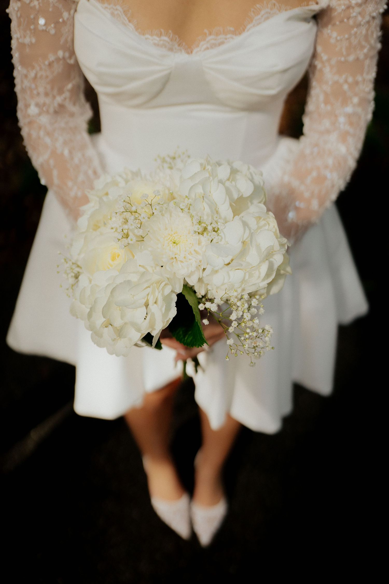A bride in a white lace dress holds an elegant bouquet of white hydrangeas against a dark background.