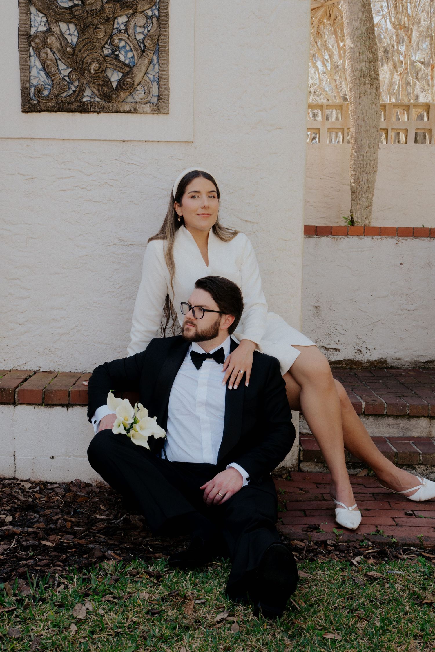 A couple in formal attire poses together for a wedding photo against a white wall with decorative artwork.